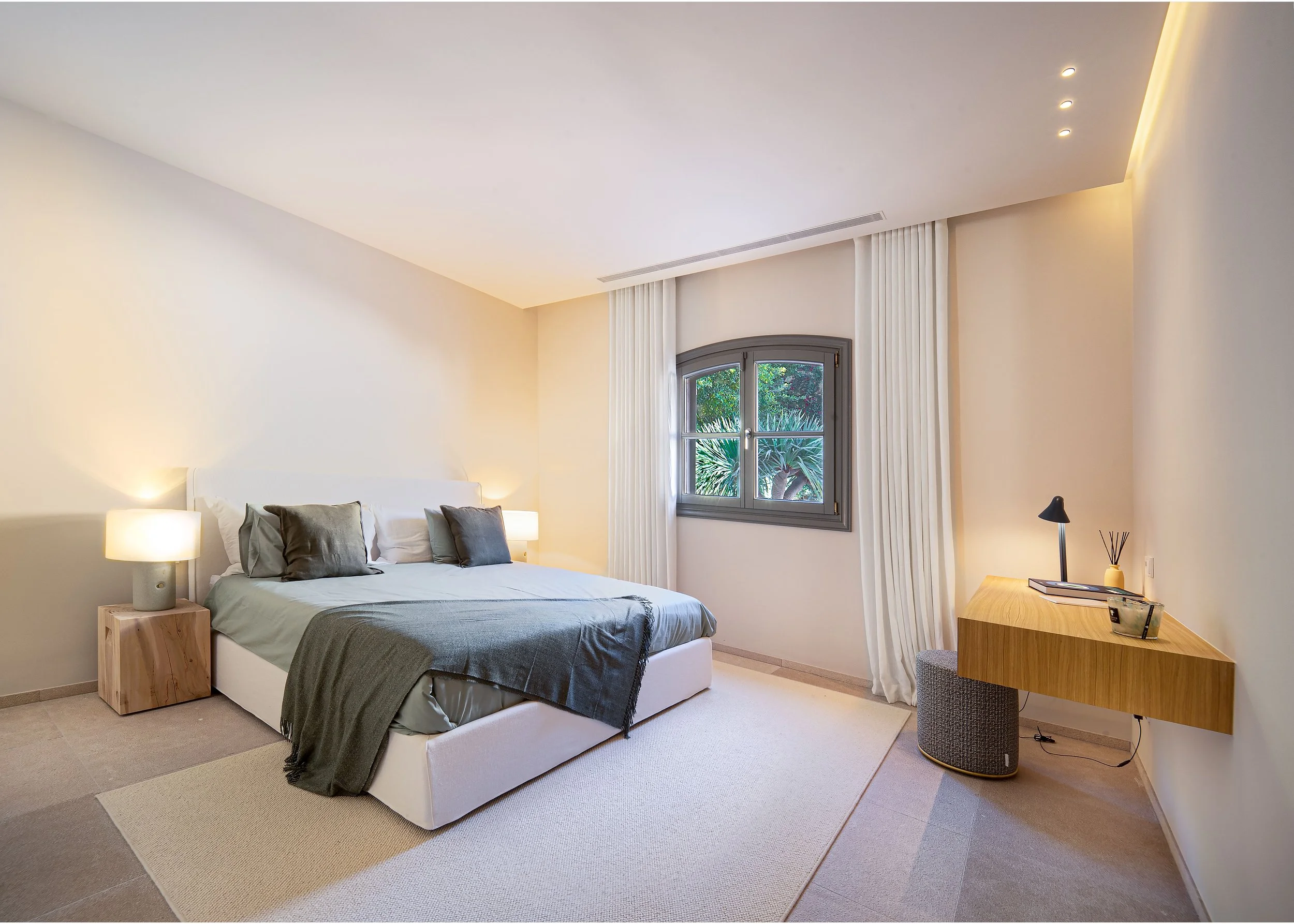 Habitación moderna con cama grande, ventanas con vista a plantas, lámparas de mesa, escritorio de madera con silla, y paredes de color claro.