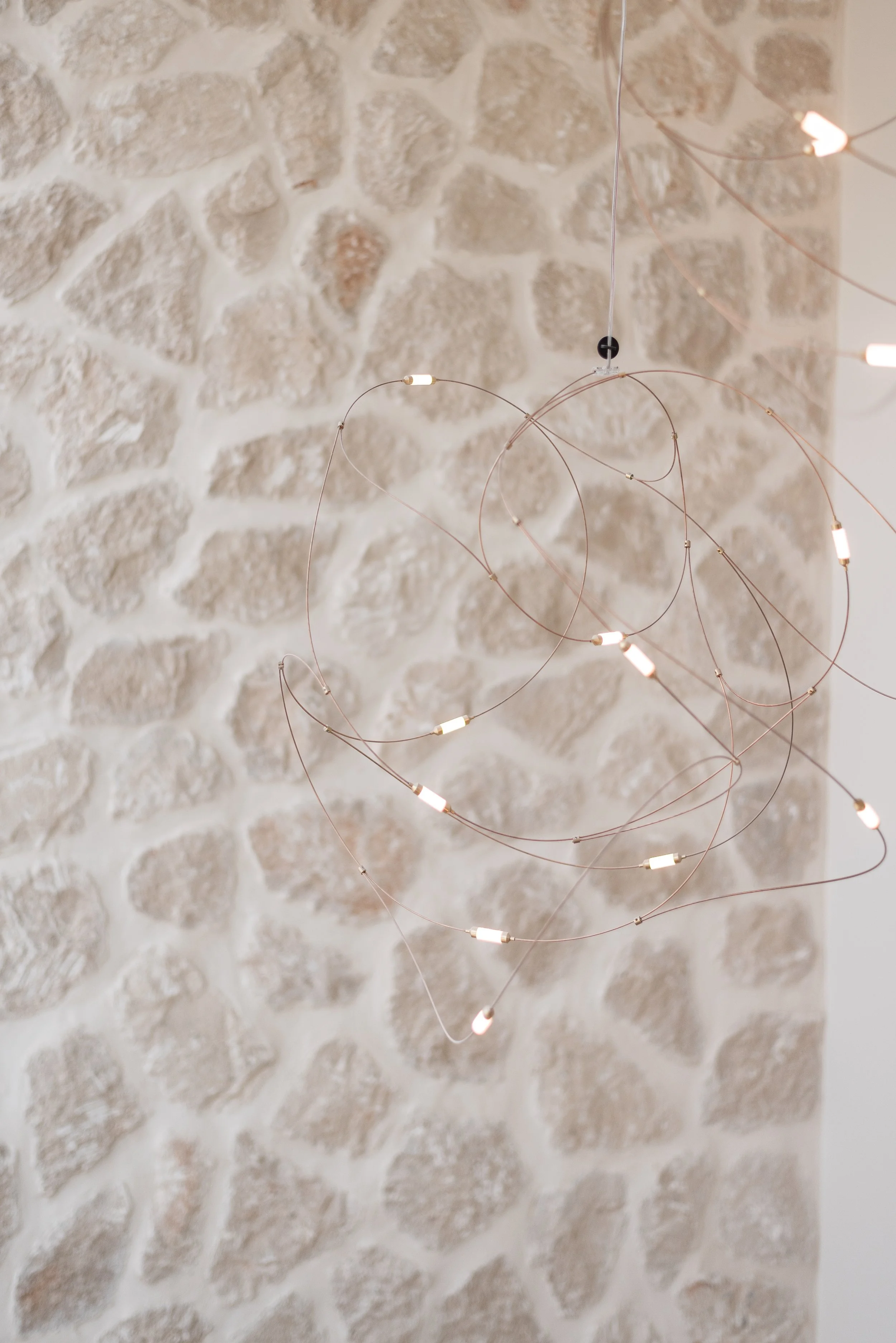Lámpara moderna suspendida con estructura de aros de cobre y luces LED, colgada frente a una pared de piedra natural beige.