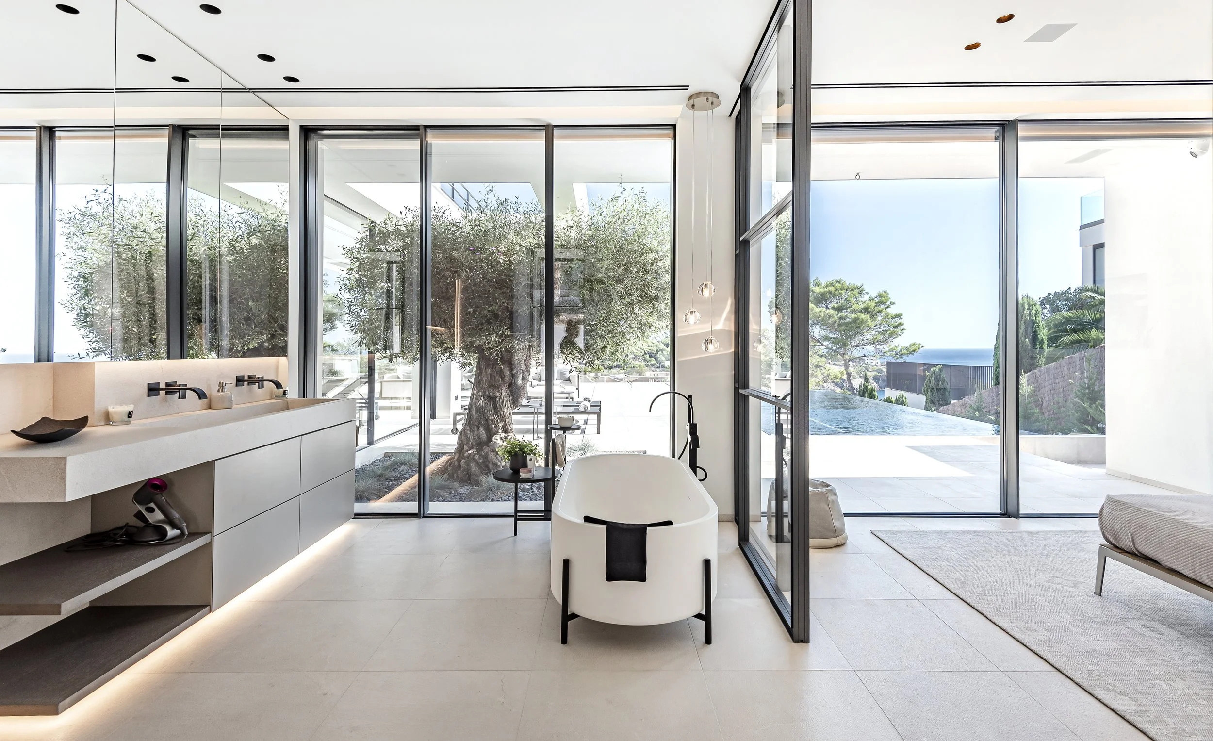 Baño moderno con bañera, vista exterior con árboles y piscina, grandes ventanas de piso a techo, luces LED y decoración minimalista.
