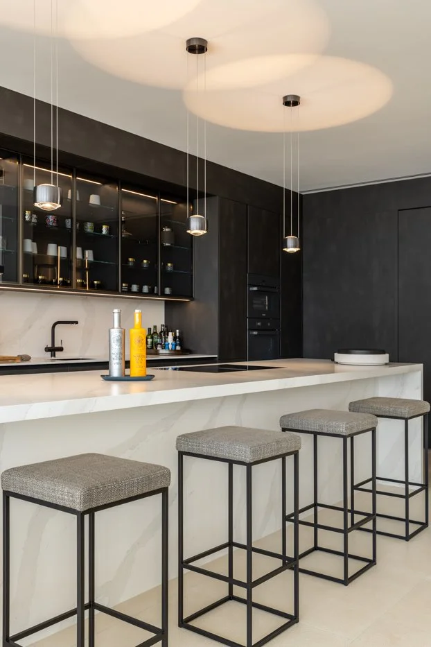 Cocina moderna con barra de granito, tres banquetas con bases de metal y asientos tapizados grises, gabinetes negros con cristaleras, y lámparas colgantes en el techo, en colores beige y negro.