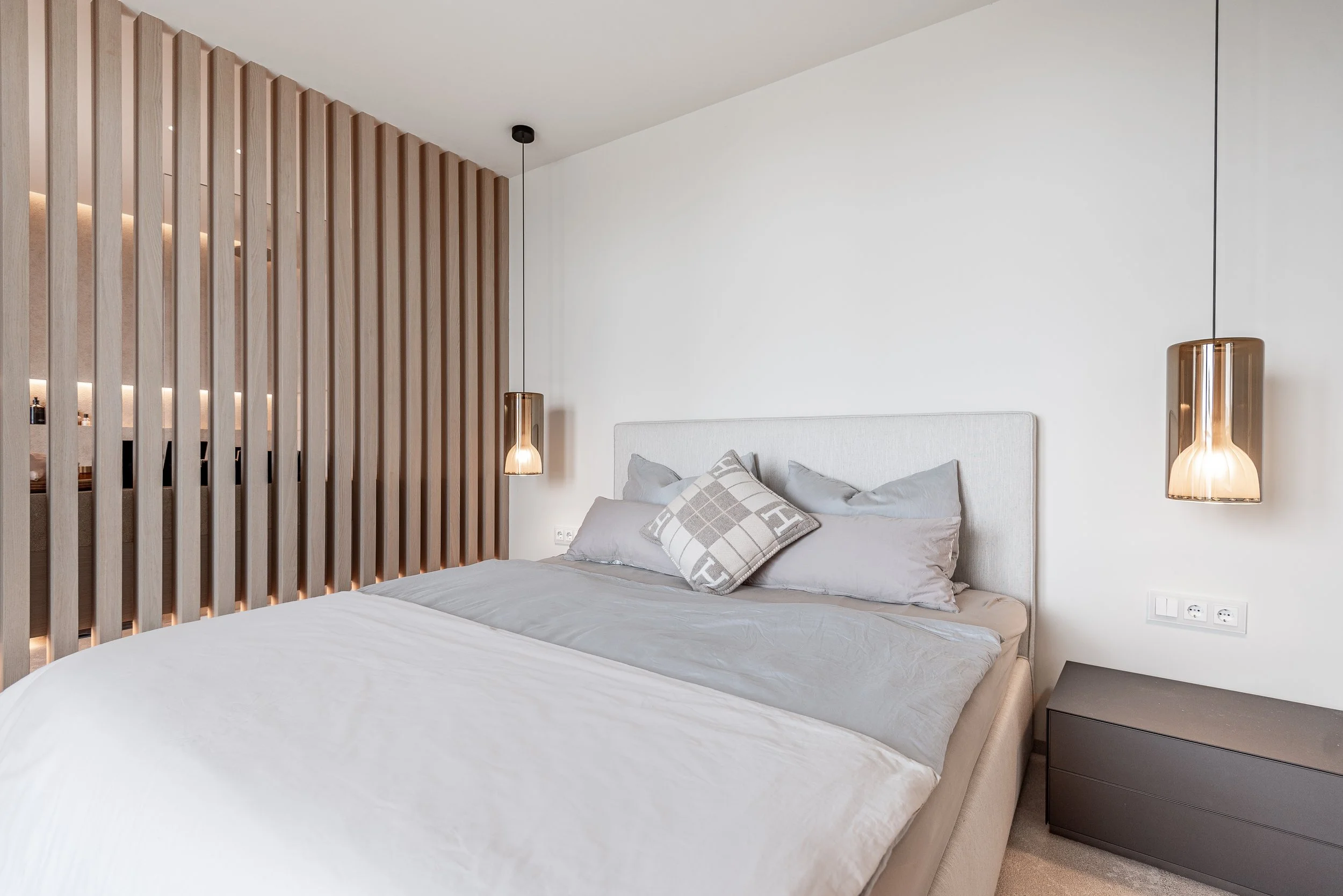 Dormitorio moderno con cabecera blanca, candelabros colgantes de vidrio en tonos dorados, pared decorativa de madera a rayas y mesa de noche gris.