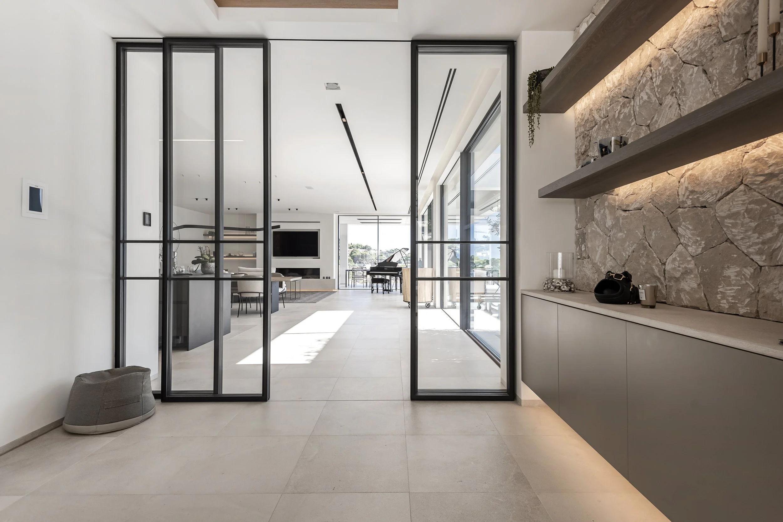 Interiores de una casa moderna con paredes blancas, puertas correderas de cristal, una sala con pizo, un piano, una pared de piedra con repisas y decoración, ventanas grandes que dejan entrar mucha luz natural.