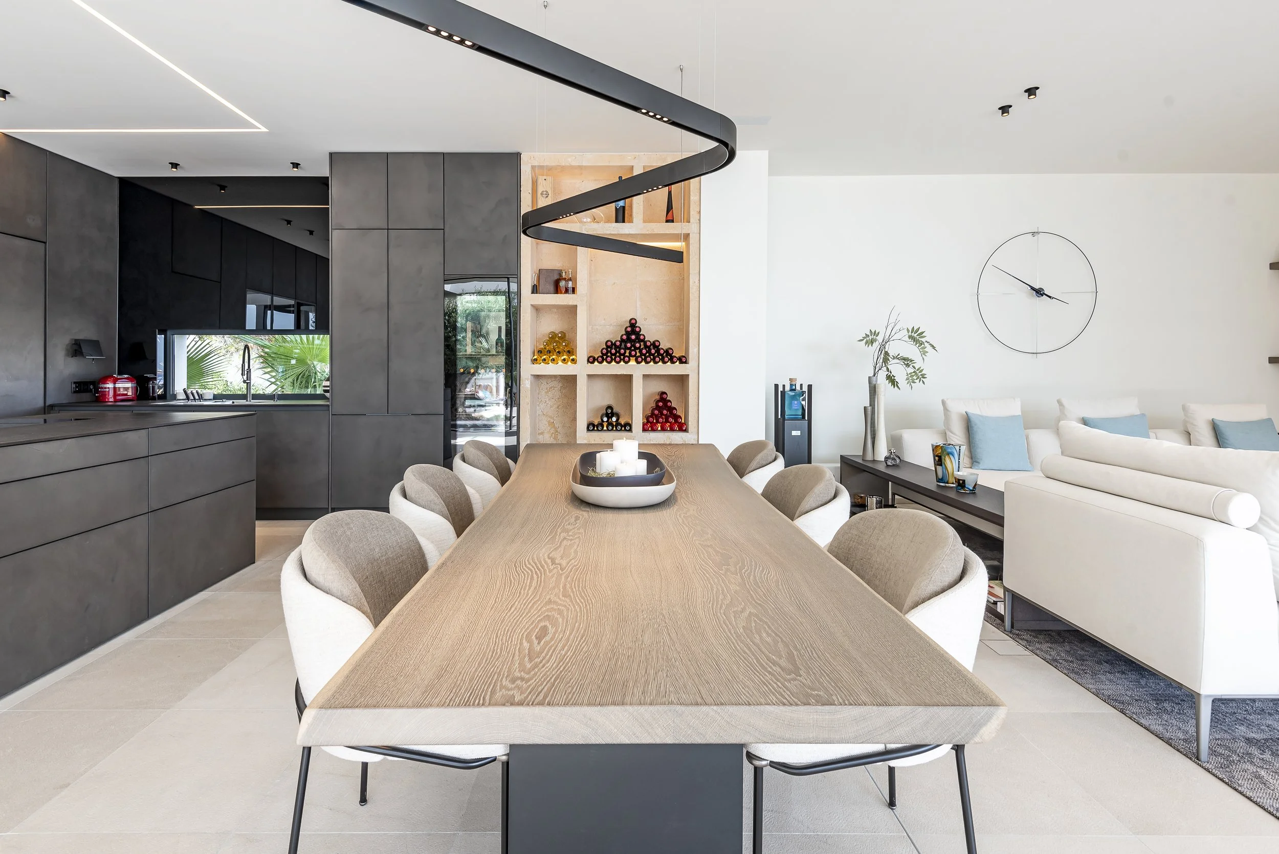 Interior moderno de sala con comedor con mesa de madera, sillas beige, sofá blanco en la sala, estantería con botellas de vino decorativo y reloj en la pared blanca.
