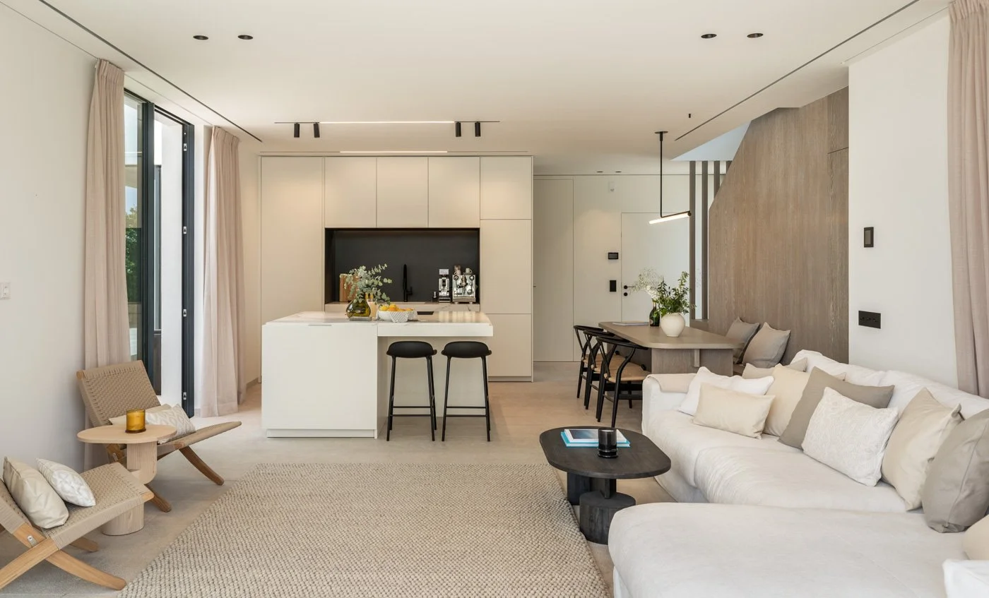 Sala de estar moderna y luminosa con sofá blanco, mesa de centro negra, orilla del sofá con libro, y sillón de madera con cojines, en el fondo cocina con barra y taburetes negros, pared de madera y plantas decorativas.