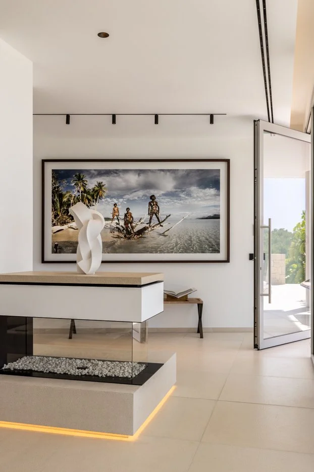 Detalle de una decoración moderna con una escultura de cerámica en una mesa y un cuadro grande de personas en un kayak en una playa tropical, en una sala con puerta de cristal abierta hacia el exterior.