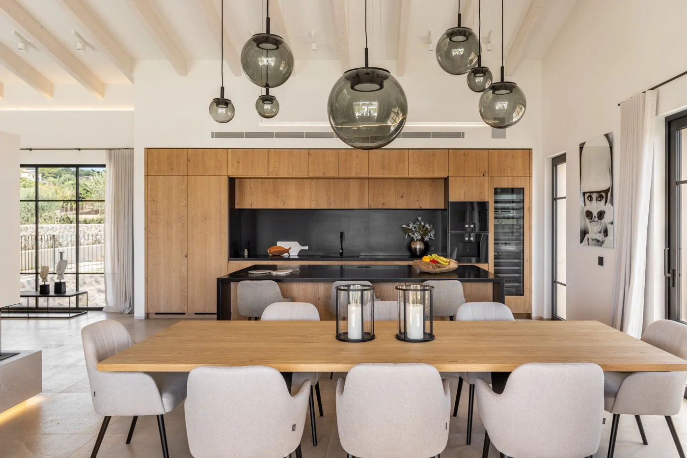 Comedor con mesa de madera y sillas blancas, cocina de fondo con gabinetes de madera y paredes negras, lámparas colgantes de burbuja, ventanas grandes con cortinas blancas, decoración minimalista y moderna.