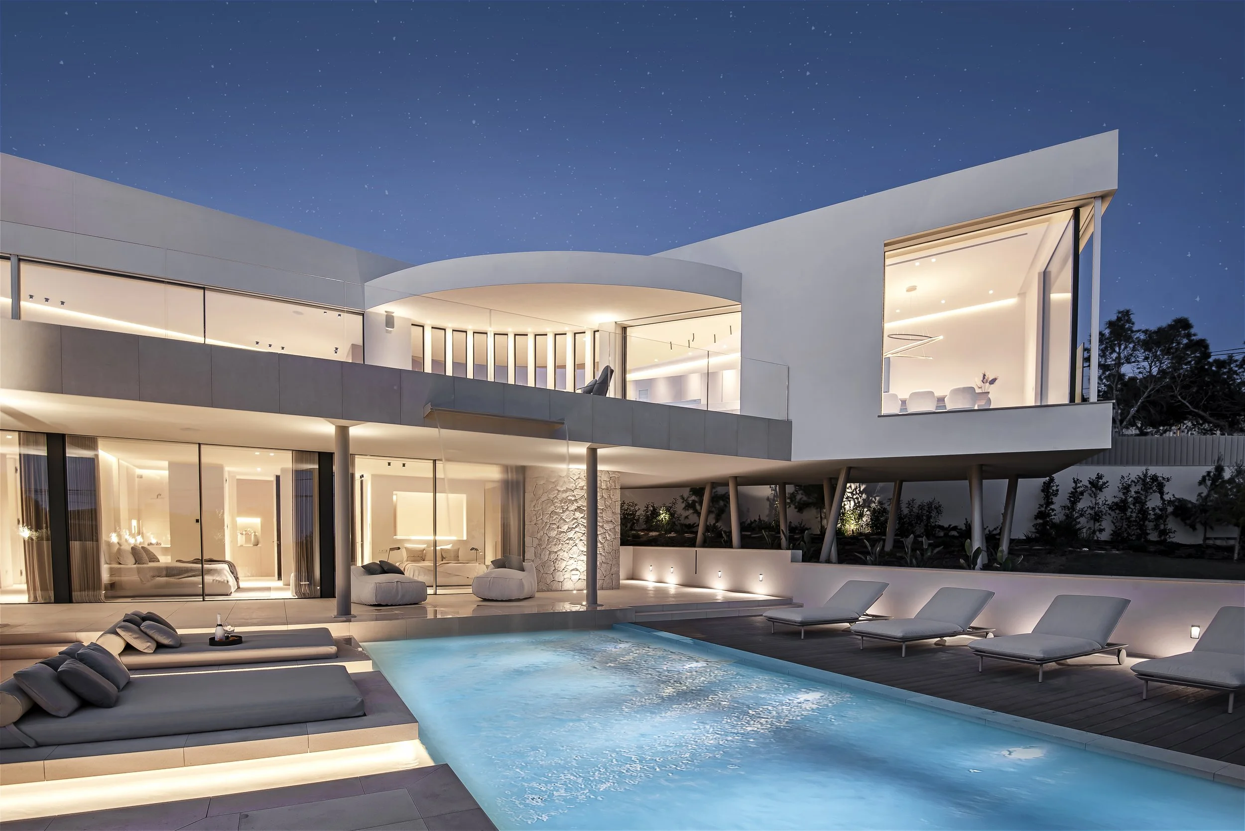 Casa moderna con iluminación nocturna, piscina y tumbonas alrededor del patio.