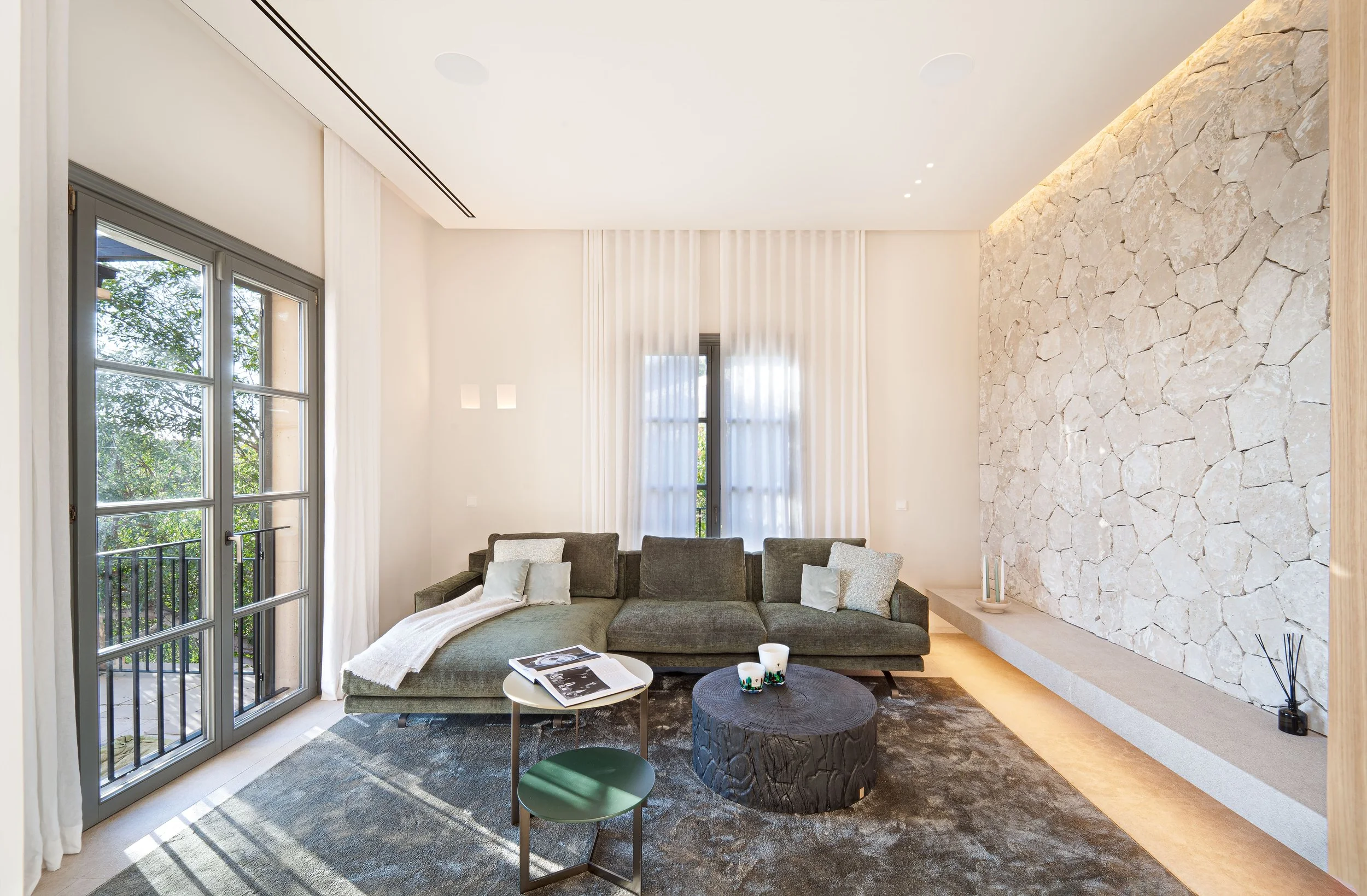 Sala de estar con muebles de tonos neutros y paredes de piedra decorativa, con ventanas grandes que dejan entrar luz natural.