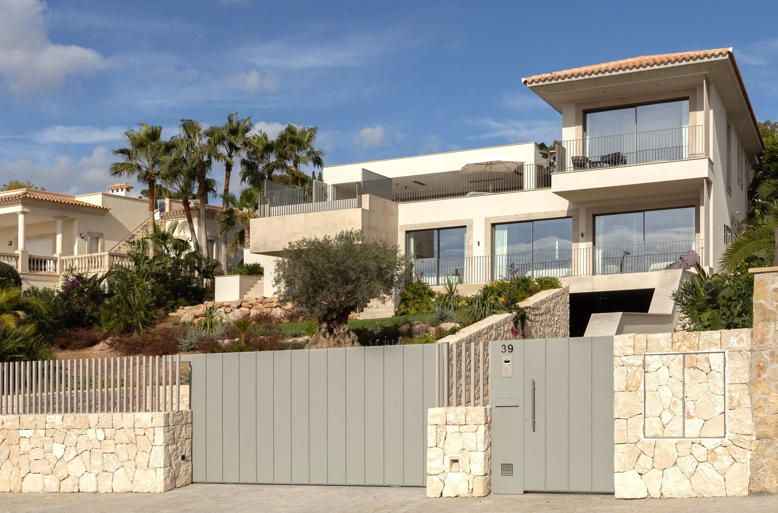 Casa moderna con múltiples pisos, terrazas y balcones, rodeada de palmeras y vegetación, con un muro de piedra en el frente y un portón de metal gris.