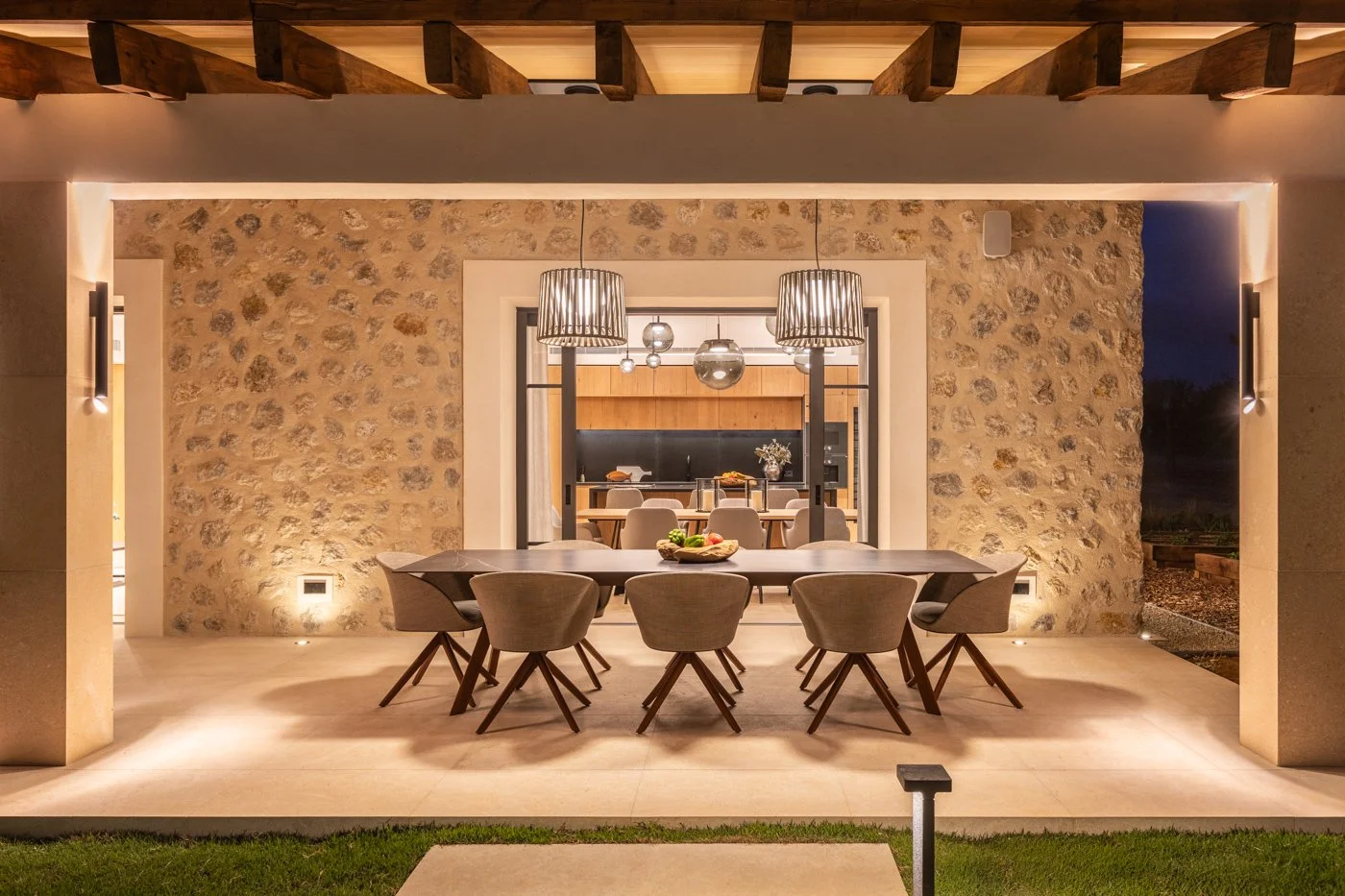 Comedor al aire libre con mesa de madera y sillas tapizadas, luces colgantes y una pared de piedra, vista a la cocina en el interior, atardecer o noche.