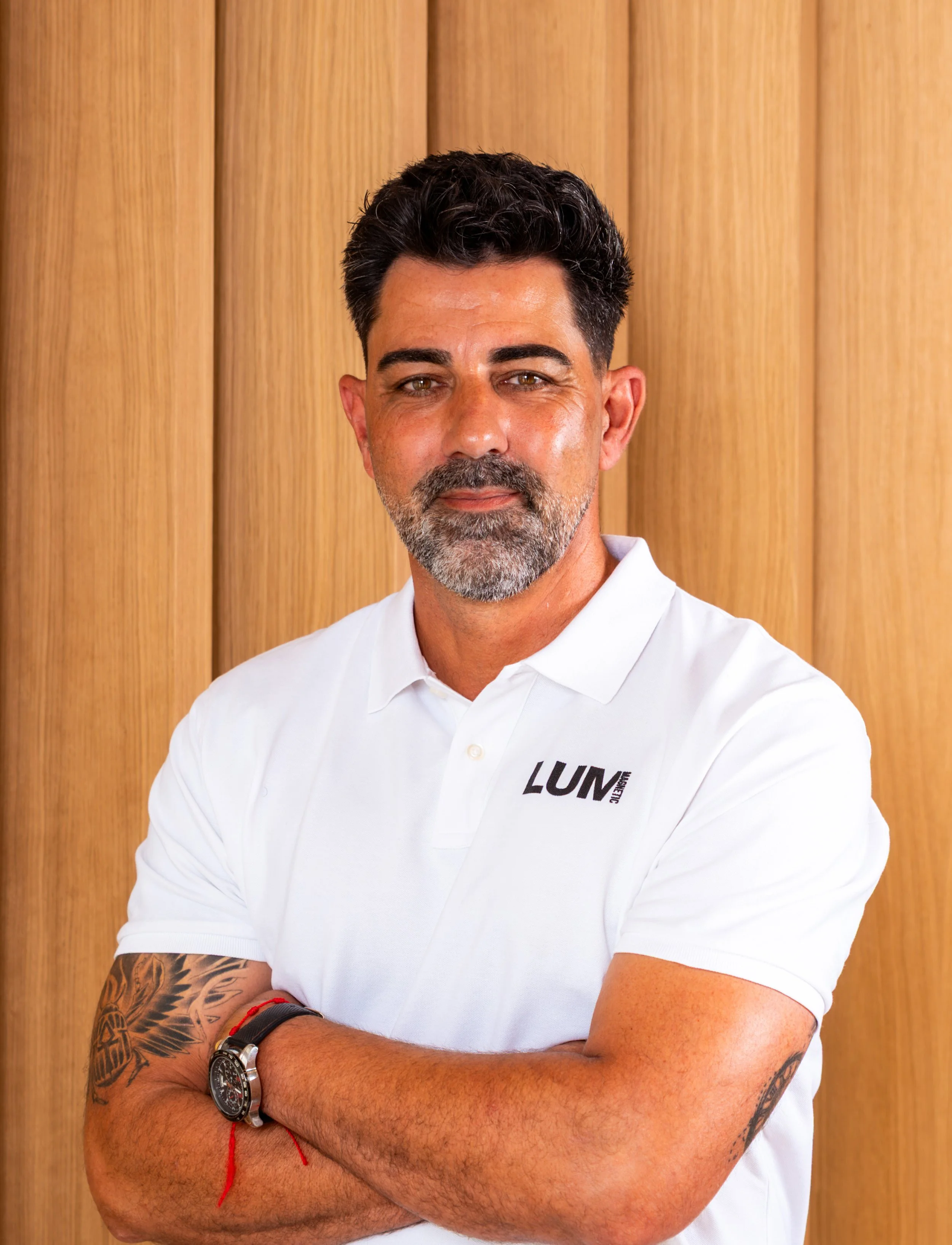 Hombre con barba y cabello oscuro, usando camiseta blanca con texto en el pecho, parando con brazos cruzados, mostrando tatuajes y reloj, fondo de madera.
