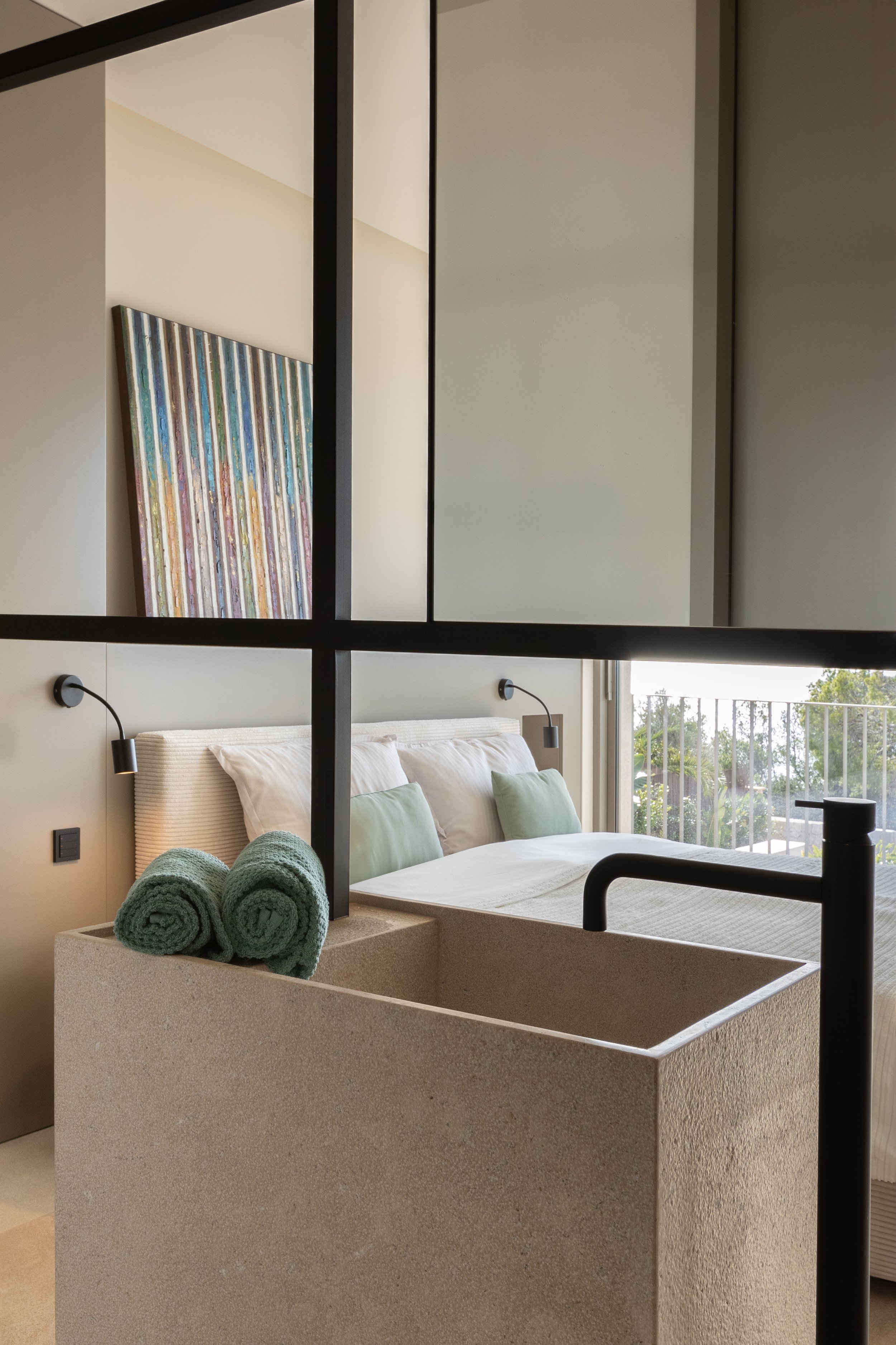 Habitación moderna con cama, almohadas, ropa de cama blanca, una bañera de piedra y toallas verdes enrolladas.