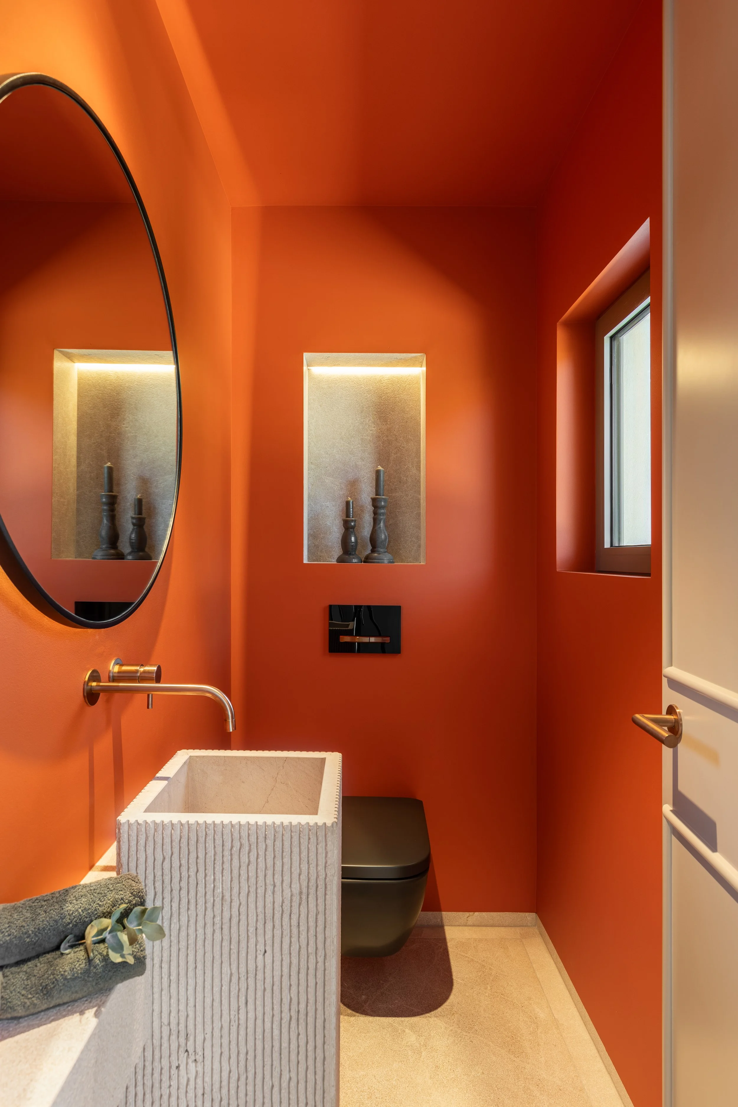 Interior de baño con paredes de color naranja, lavabo de piedra, espejo redondo, ventana, y accesorios de decoración y funcionales.