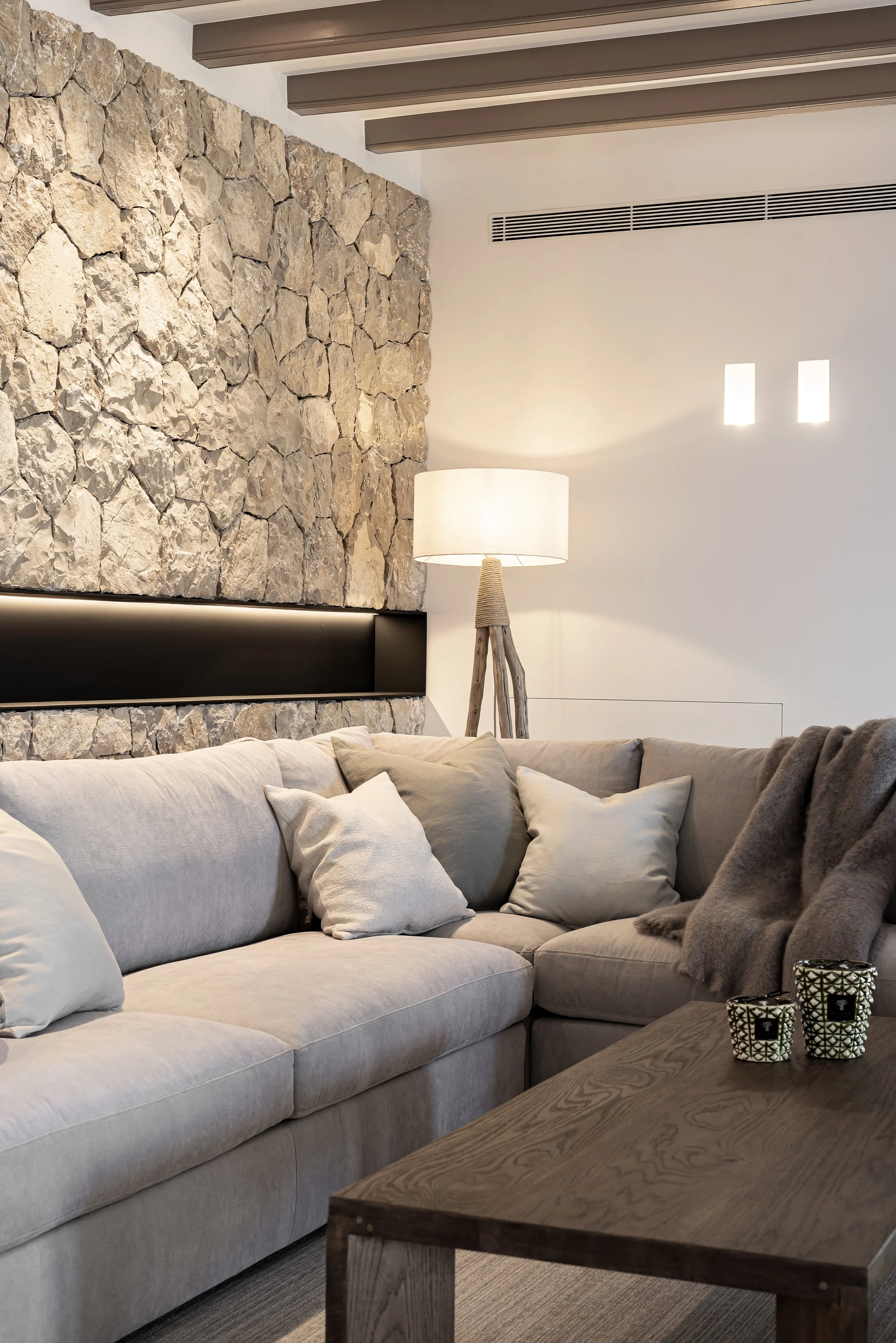 Sala de estar con sofá beige, cojines blancos y oscuros, lámpara de pie de madera, pared de piedra y pared blanca con dos ventanas pequeñas y un conducto de ventilación.