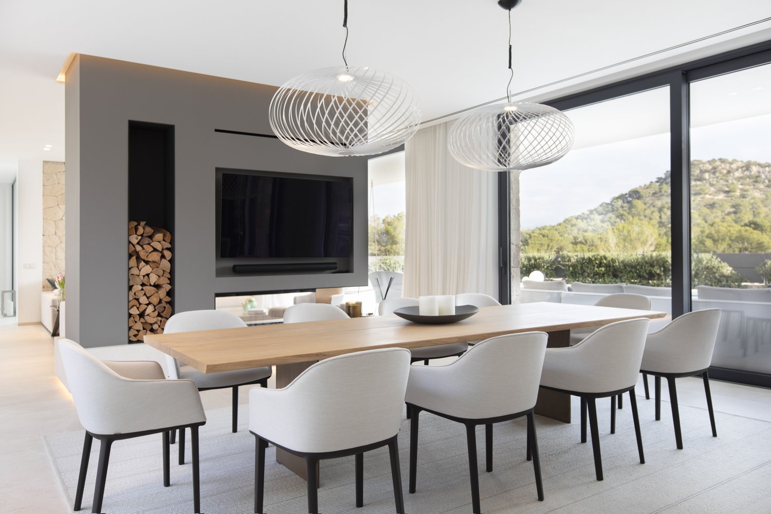 Comedor moderno con mesa de madera y sillas blancas, grandes ventanas con vista a un paisaje natural, chimenea con leña apilada y televisión en la pared, decoraciones minimalistas y lámparas elegantes colgantes.