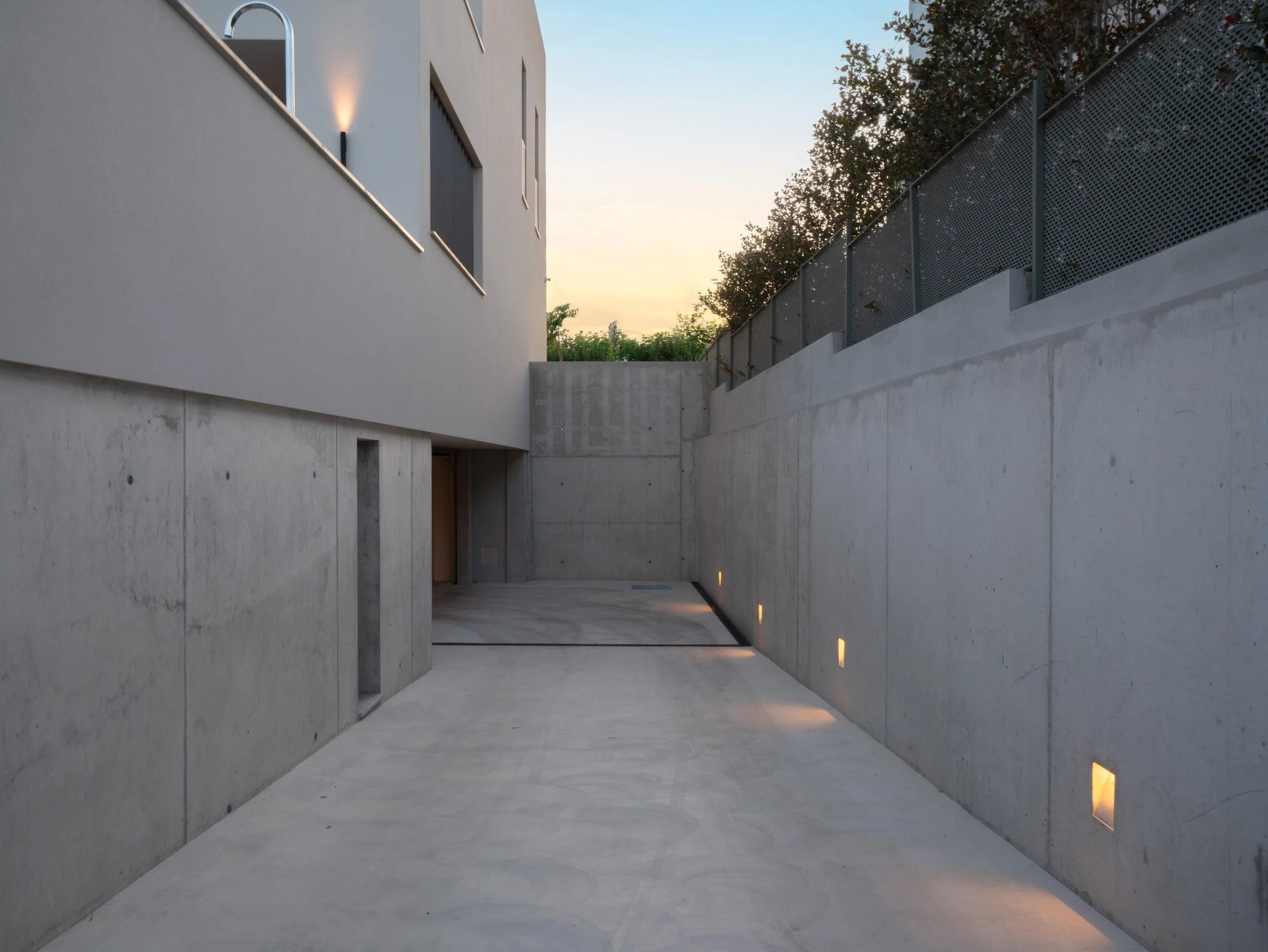 Pasillo abierto con paredes de concreto y luces empotradas en el suelo, con un muro de metal y árboles al fondo al atardecer.