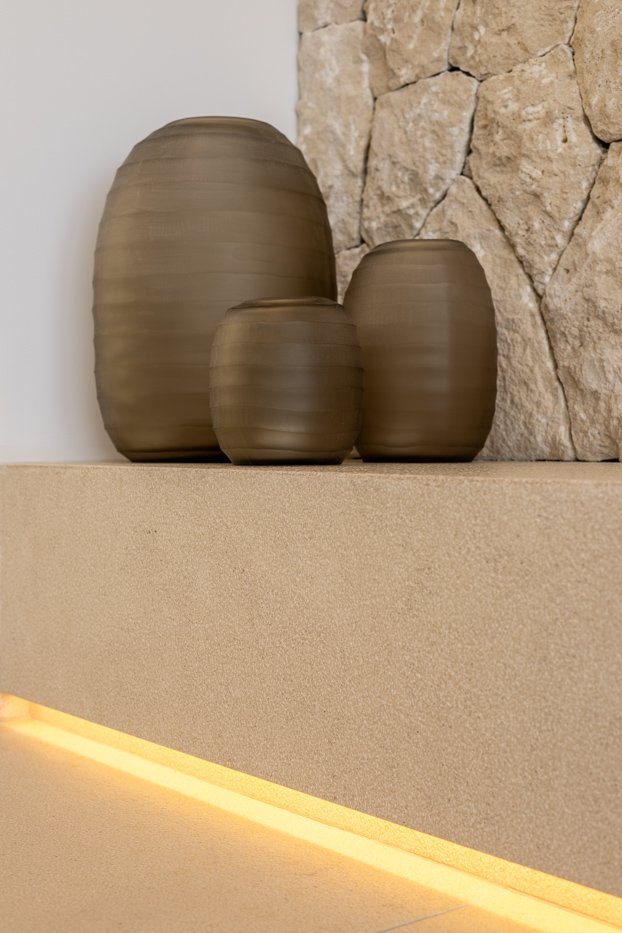 Tres jarrones de cerámica en tonos beige sobre una repisa de color crema, con una pared de piedra en el fondo.