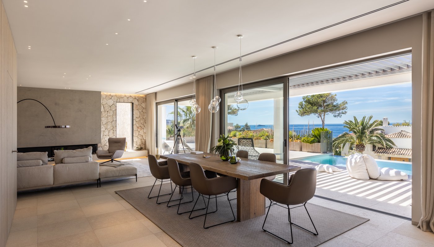 Casa moderna con vista al mar, sala de estar y comedor con muebles elegantes, piscina y paisaje tropical con palmeras.