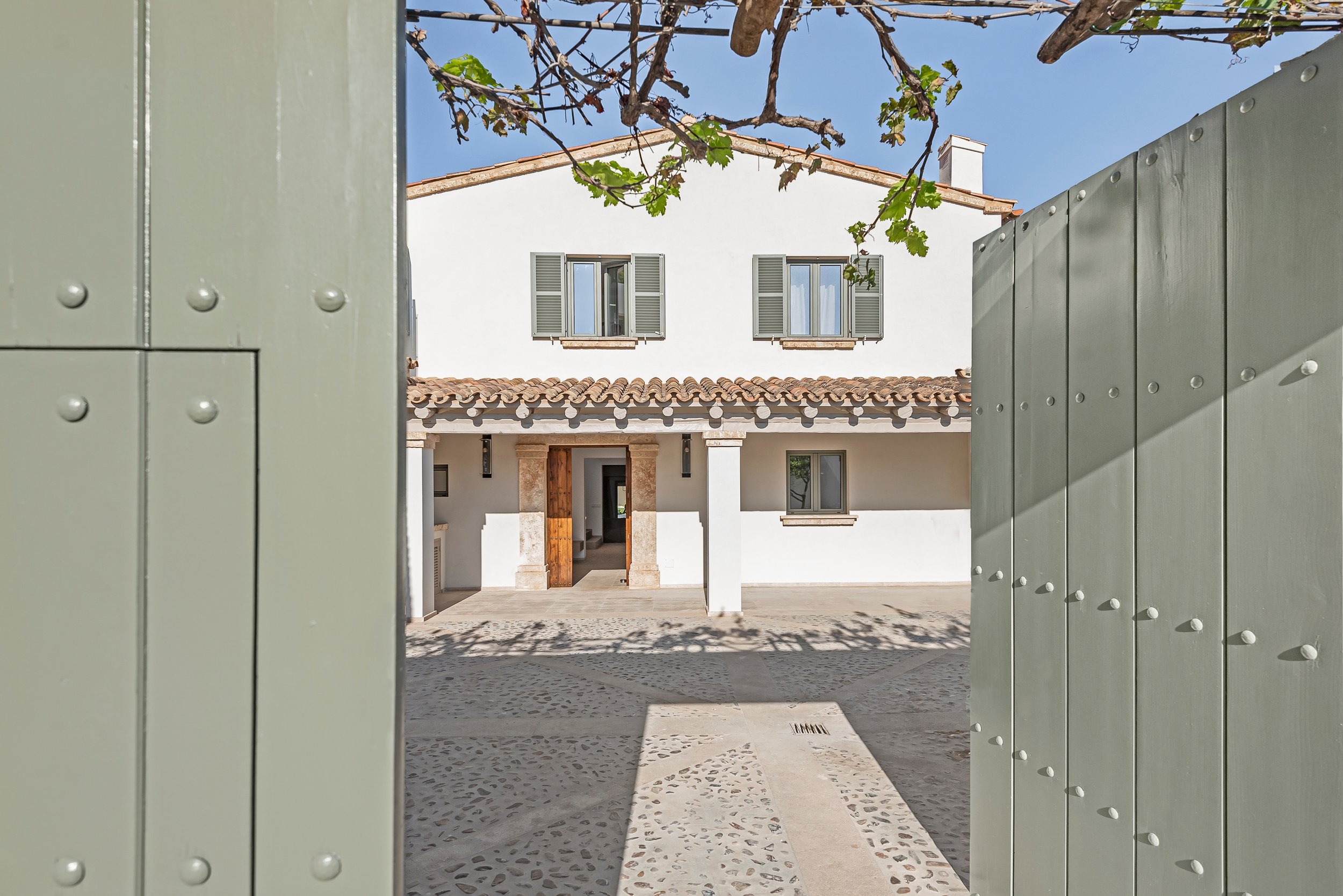 Vista de una casa moderna desde el umbral de una puerta verde gigante, con paredes blancas, ventanas con persianas grises y un tejado de tejas, bajo un cielo azul y ramas de árbol en primer plano.