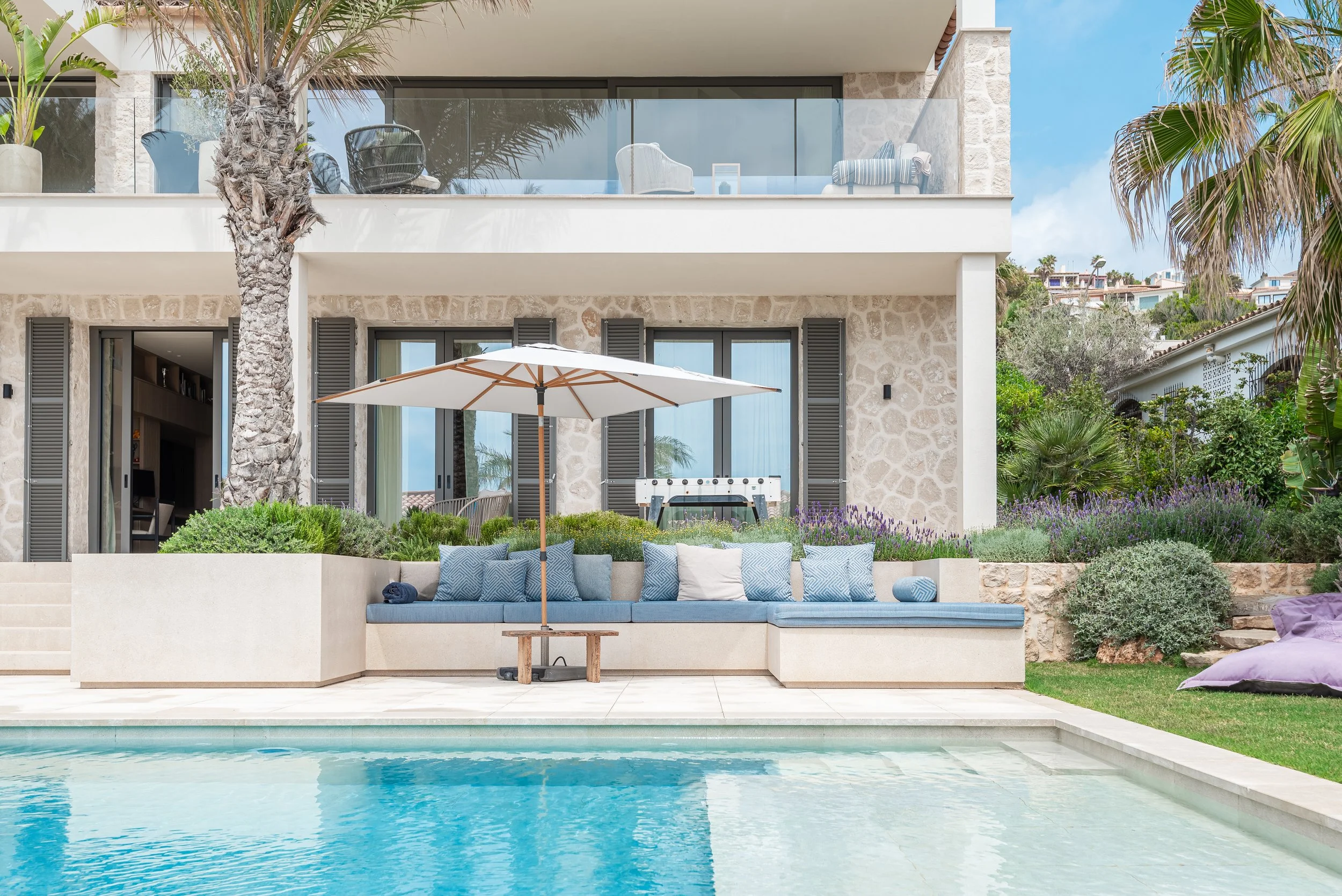 Vista de una casa moderna con balcón en un entorno tropical, con palmeras, vegetación y una piscina en primer plano.
