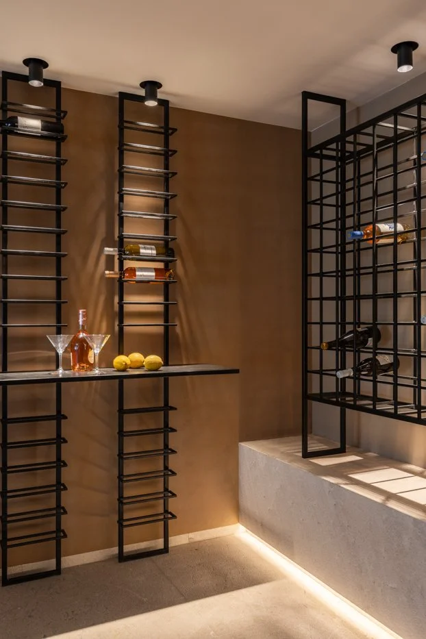 Estanterías negras con botellas de vino en un espacio moderno con paredes marrones y suelo de piedra, iluminación tenue y una mesa con copas y limones.