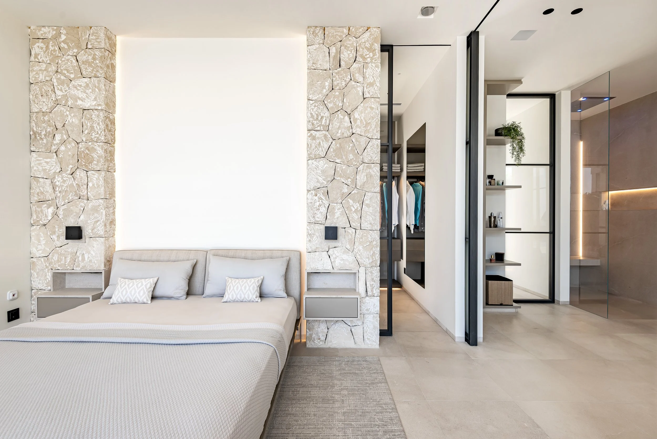 Habitación moderna con cama grande, paredes de piedra y estanterías abiertas con ropa y decoraciones, división de vidrio y estudio luminoso