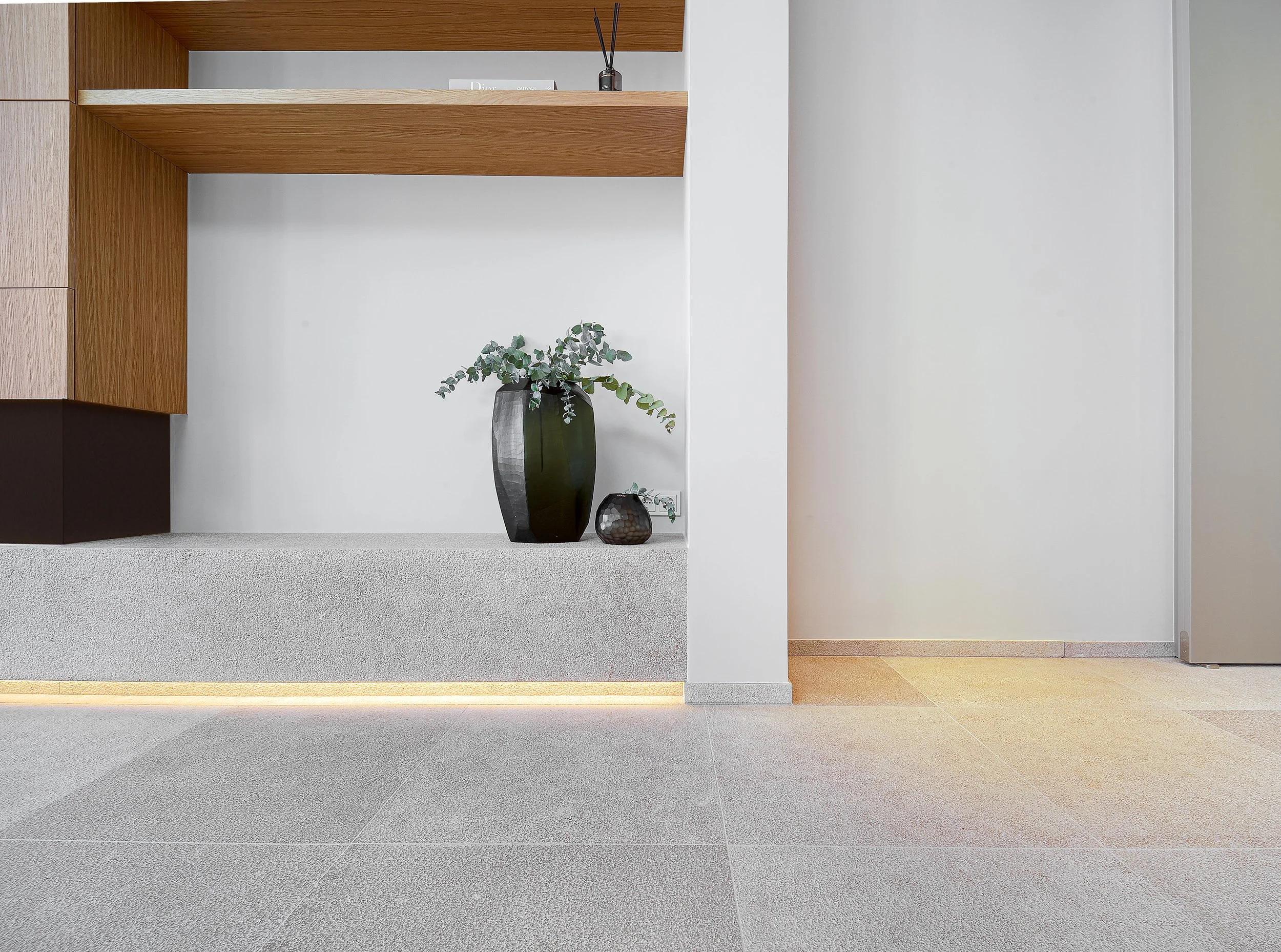 Interior de una sala moderna con suelo de baldosas grises, pared blanca y decoraciones minimalistas, incluyendo una planta en un jarrón negro y una pequeña escultura en la mesa.