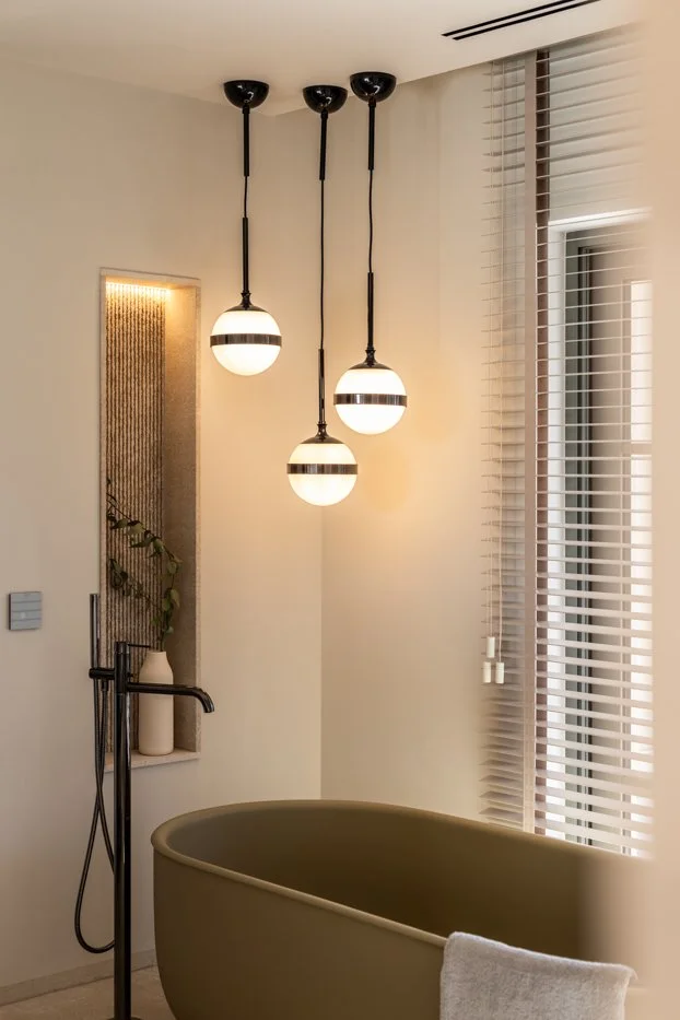 Tres lámparas colgantes con bolas blancas y detalles oscuros iluminan una bañera en un baño moderno con cortinas de persiana y detalles decorativos en la pared.
