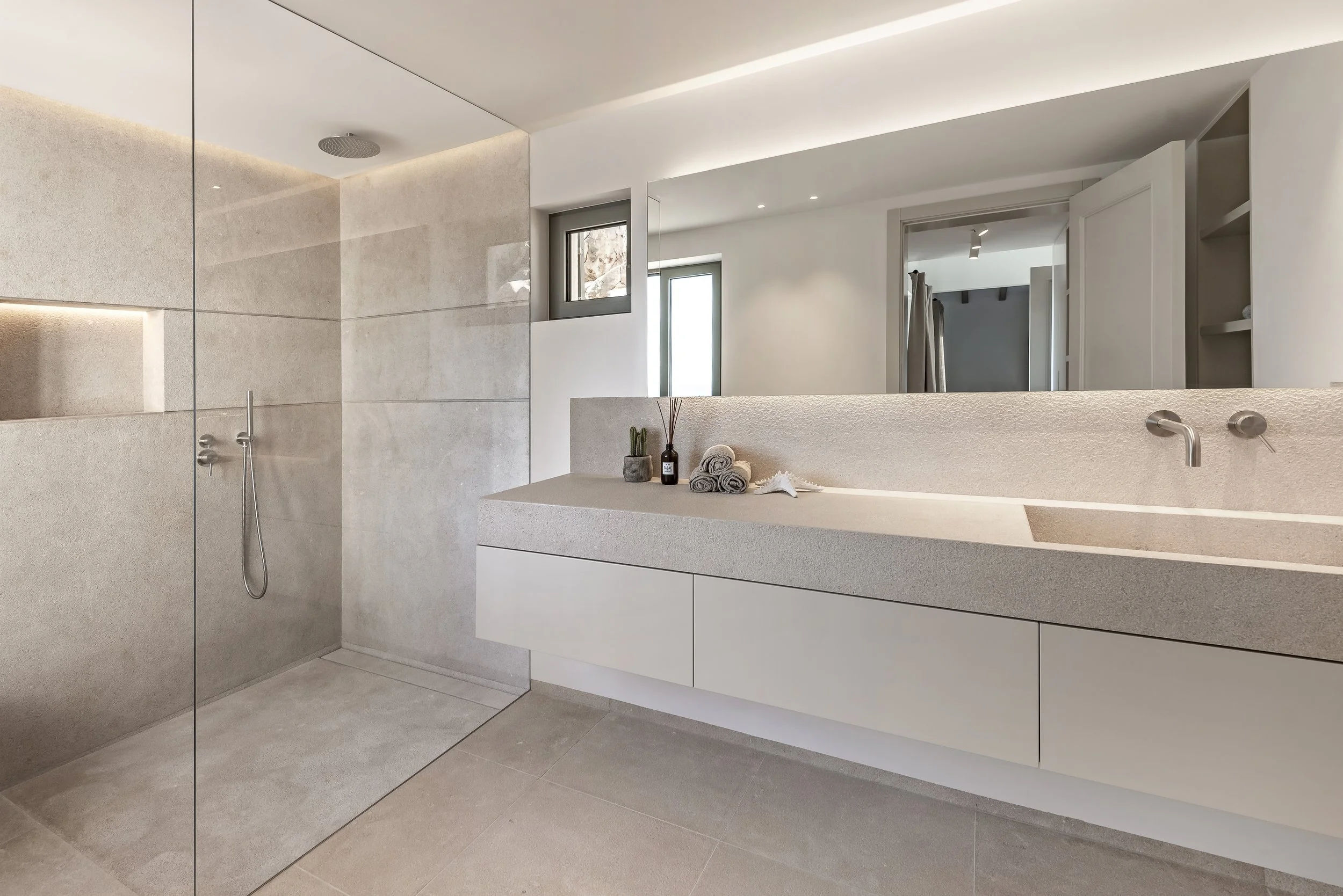 Baño moderno con paredes y suelo de mármol beige, ducha sin puerta, accesorios de baño minimalistas, ventana y espejo grande.
