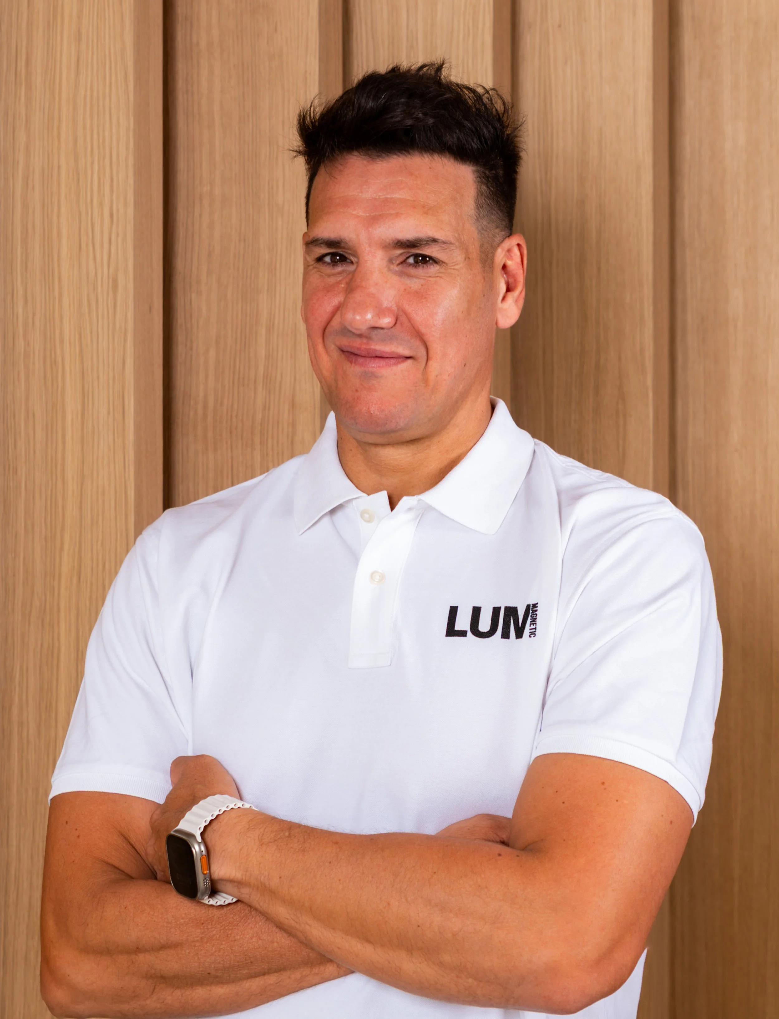 Retrato de un hombre con peinado oscuro y barba leve, vestido con una camiseta blanca con logotipo en el pecho, de piel morena clara, con brazo cruzado y mostrando un reloj inteligente, posando frente a una pared de madera.