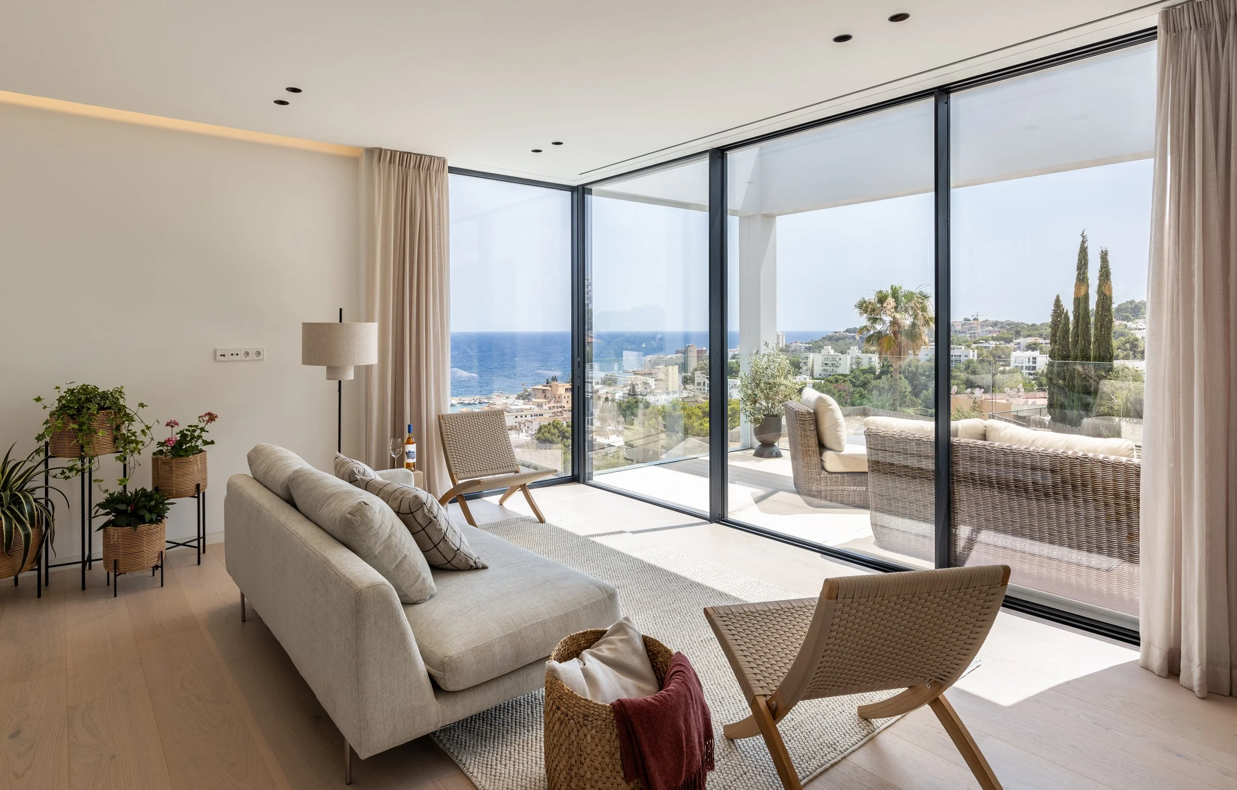 Salón con sofá beige, plantas, y vista panorámica al mar y la ciudad a través de ventanas de piso a techo.