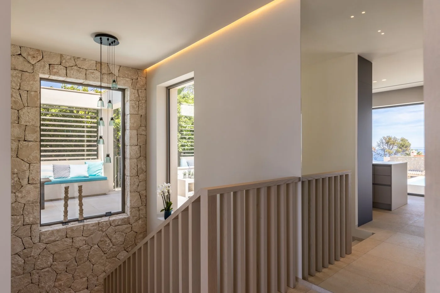Vista interior de una casa moderna con paredes de piedra y ventanas grandes que dejan entrar luz natural, balcón con almohadones y vista al exterior con árboles.