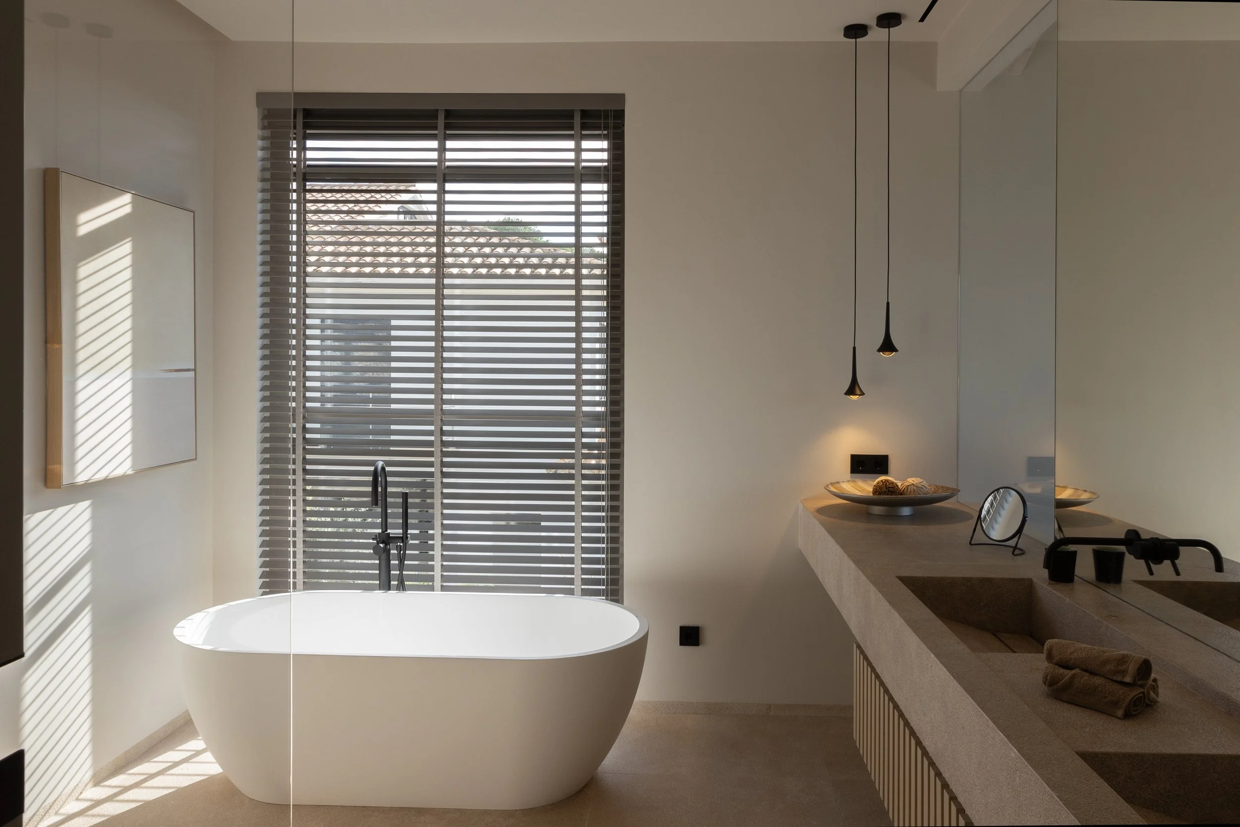 Baño moderno con bañera de pie, ventana con persianas, iluminación suspendida y encimera con varios artículos, toallas y espejo.