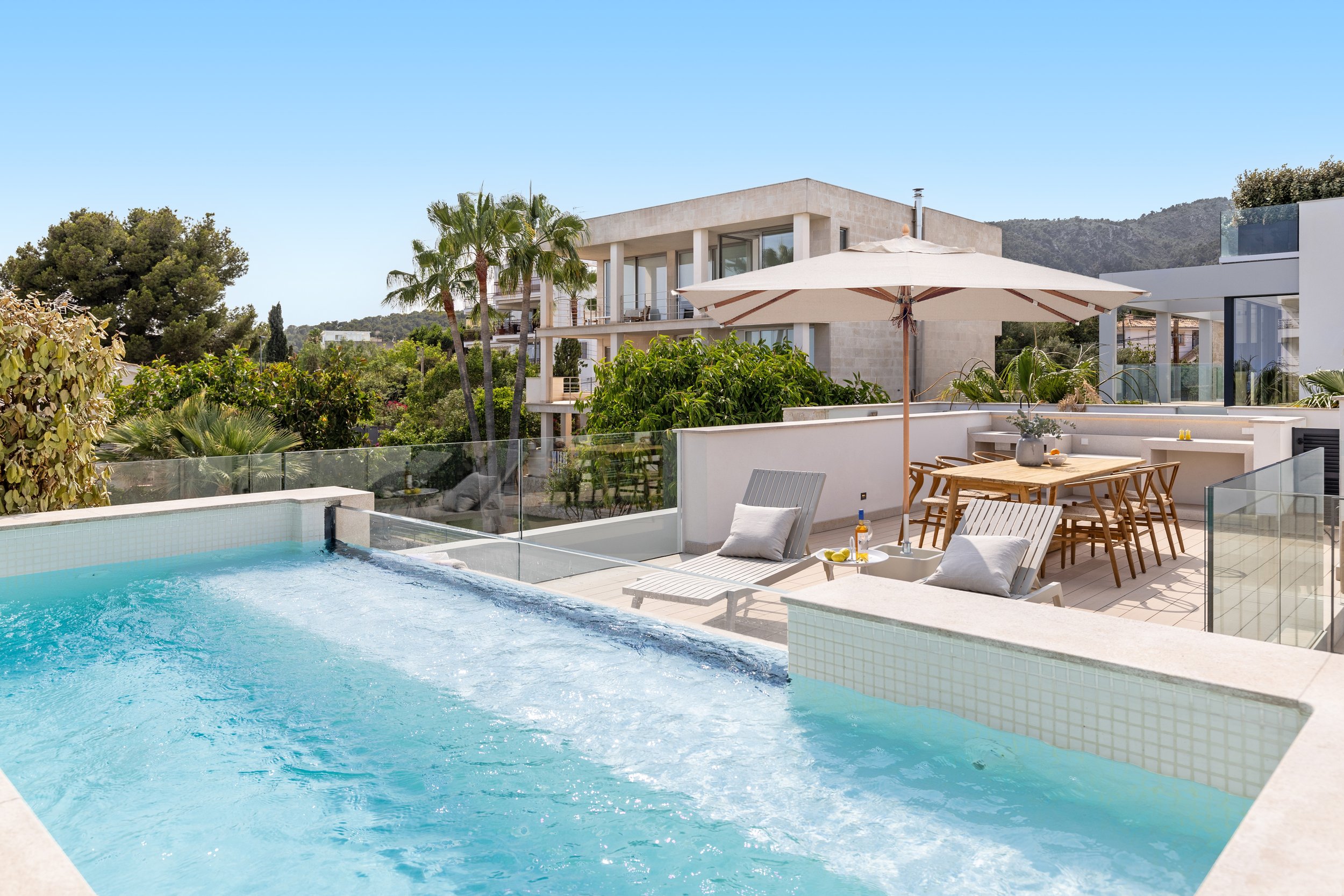 Vista de una piscina con una terraza que tiene una mesa de madera, sillas y sombrillas, con árboles y casas modernas en el fondo bajo un cielo despejado.