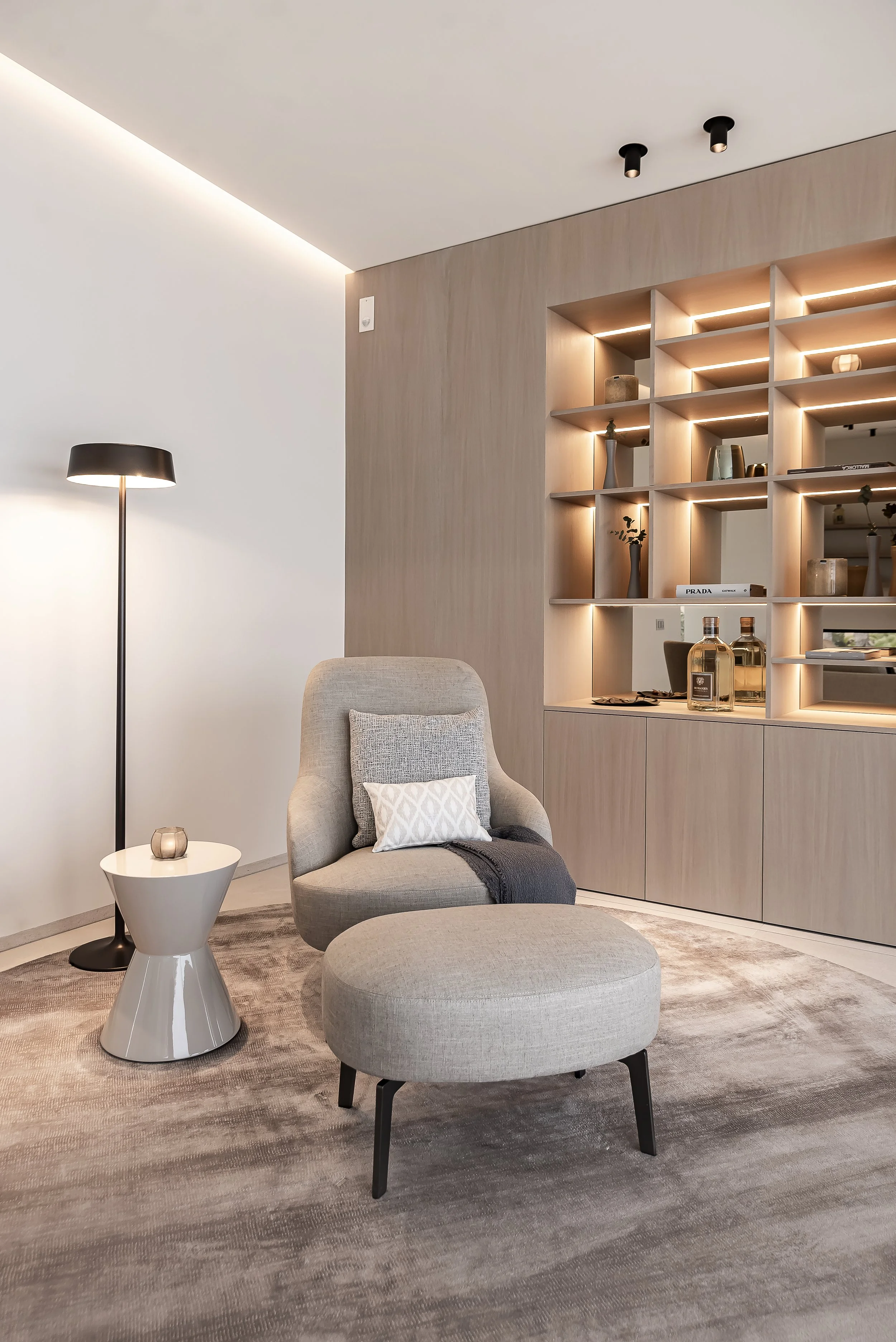Sala moderna con silla gris, mesa auxiliar y estantería con objetos decorativos, libros y plantas. Alfombra beige y luz ambiental