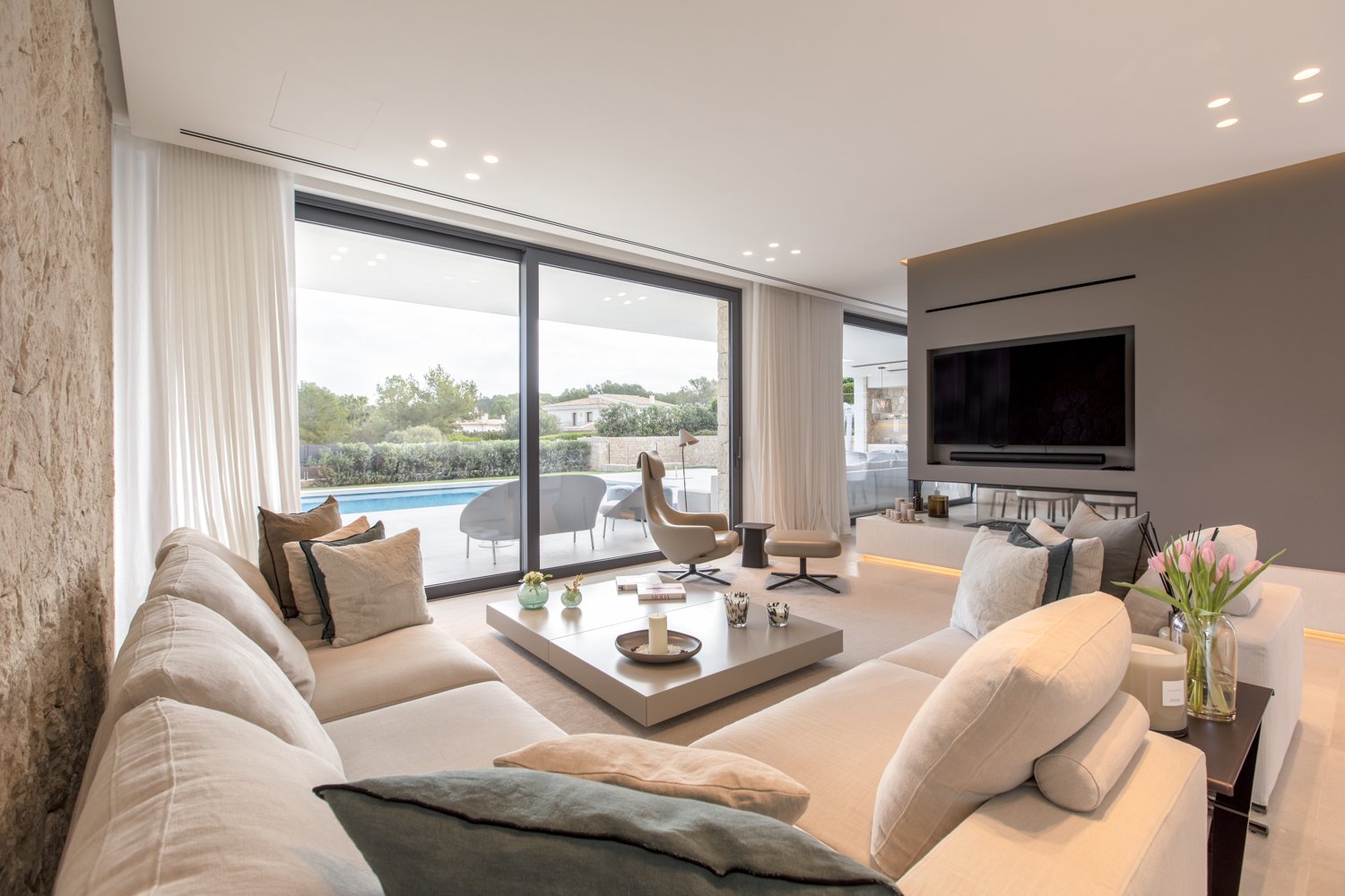 Sala moderna con sofá beige, mesa de centro, sillas de descanso, televisión en pared, puertas de cristal que dan a la piscina