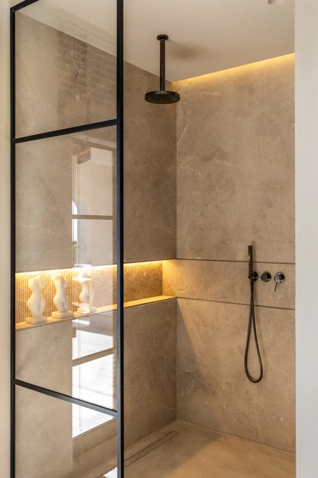 Ducha moderna con paredes de piedra beige, ducha de efecto lluvia en el techo, manguera y controles en la pared, y una repisa con esculturas y luz LED empotrada.