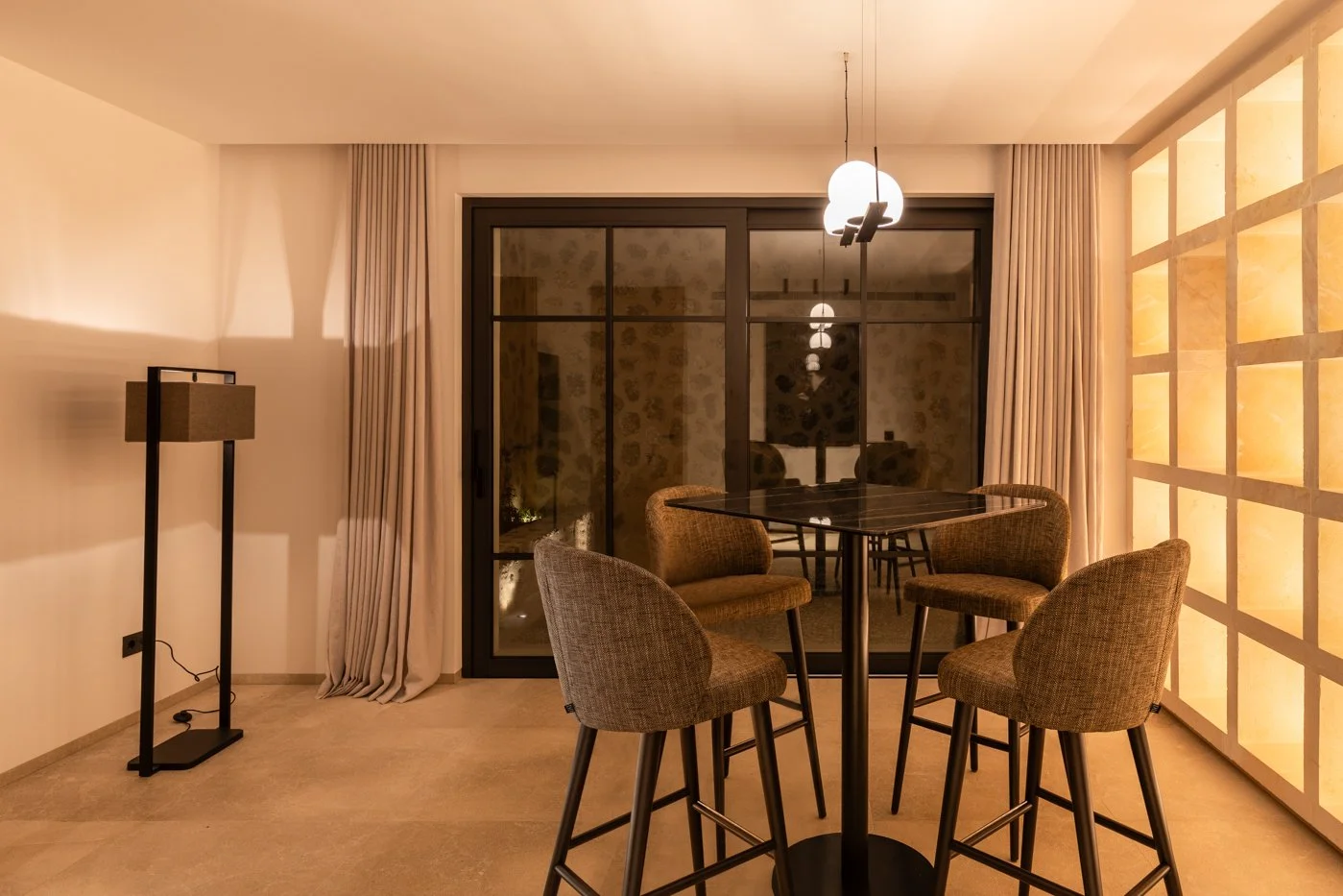 Sala con mesa alta, cuatro sillas tapizadas, cortinas beige, lámpara de techo y pared de bloques de luz
