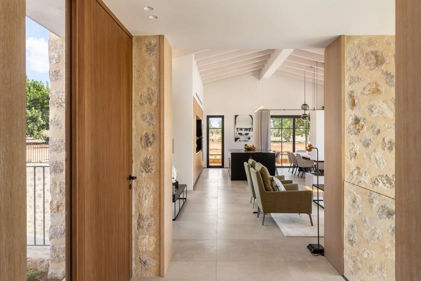 Vista del interior de una casa moderna con paredes de piedra y madera, sala con sillones verdes y comedor con sillas blancas, grandes ventanas y puerta con vista al exterior