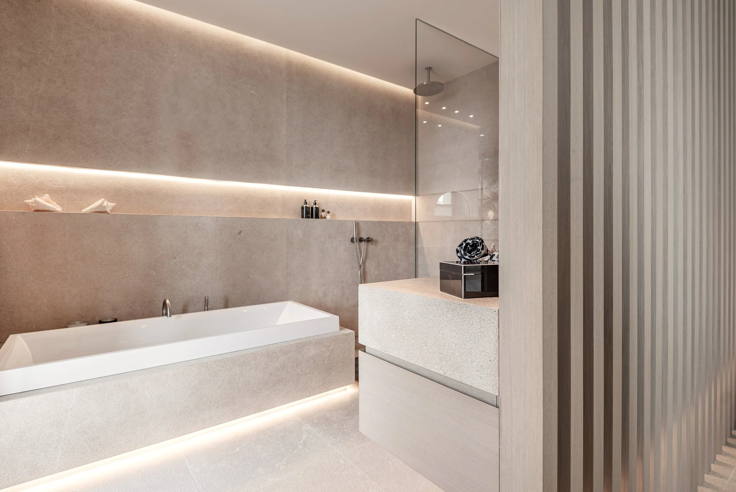 Baño moderno con bañera, muro de piedra beige, paredes de vidrio y techo con luces empotradas.