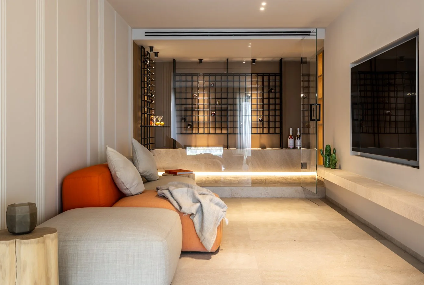 Sala moderna con sofá gris y naranja, televisión en la pared, decoraciones verdes y una pared de botellas de vino en el fondo.