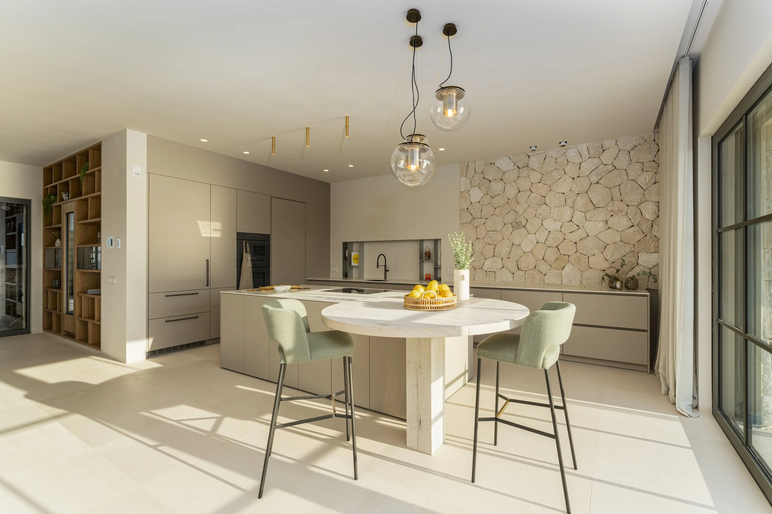Cocina moderna con isla central blanca, mesa redonda con cuatro sillas verdes, paredes de piedra, estanterías abiertas y gran ventana o puerta de cristal.