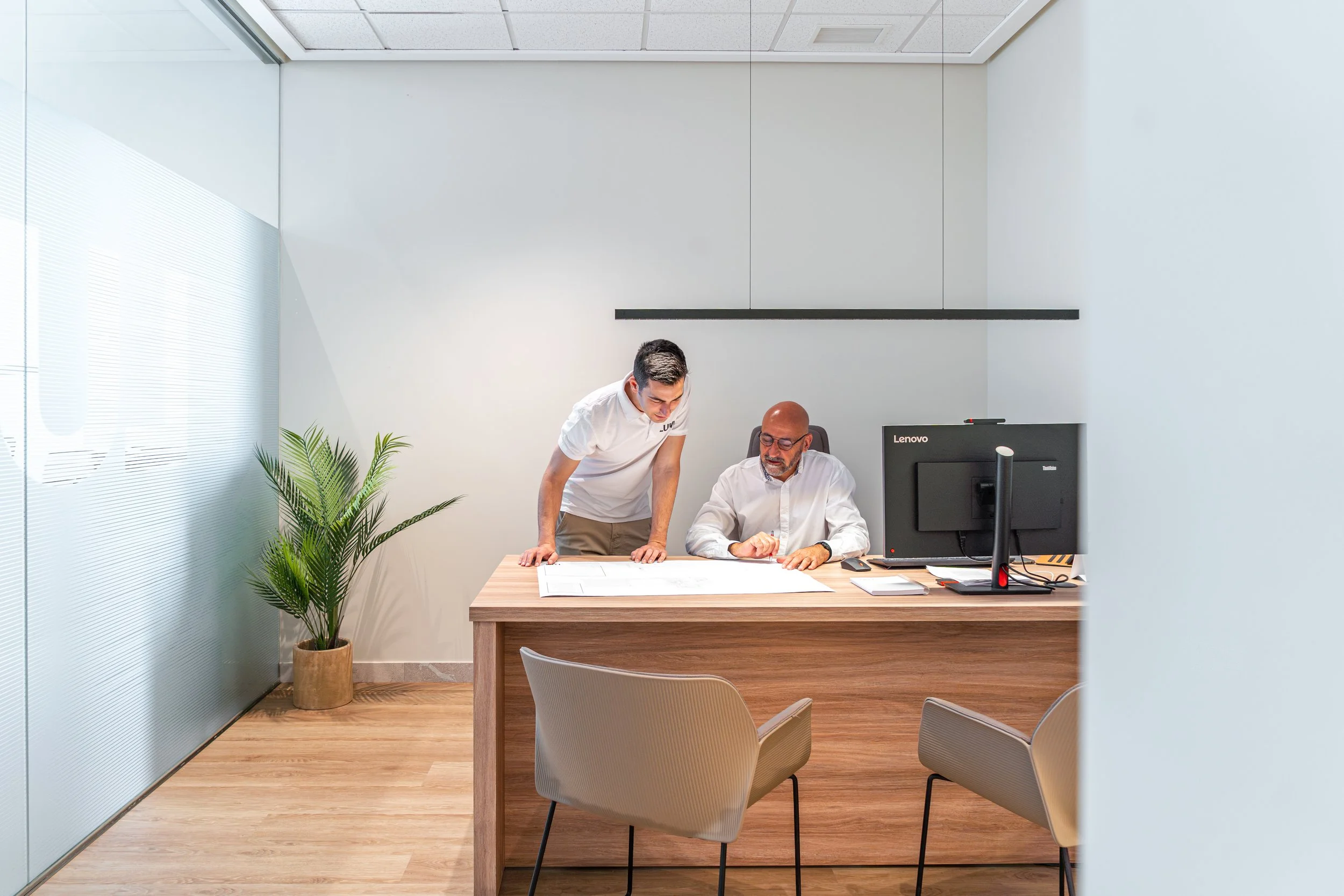 Dos hombres en una oficina revisando documentos sobre una mesa, con una computadora y plantas decorativas.