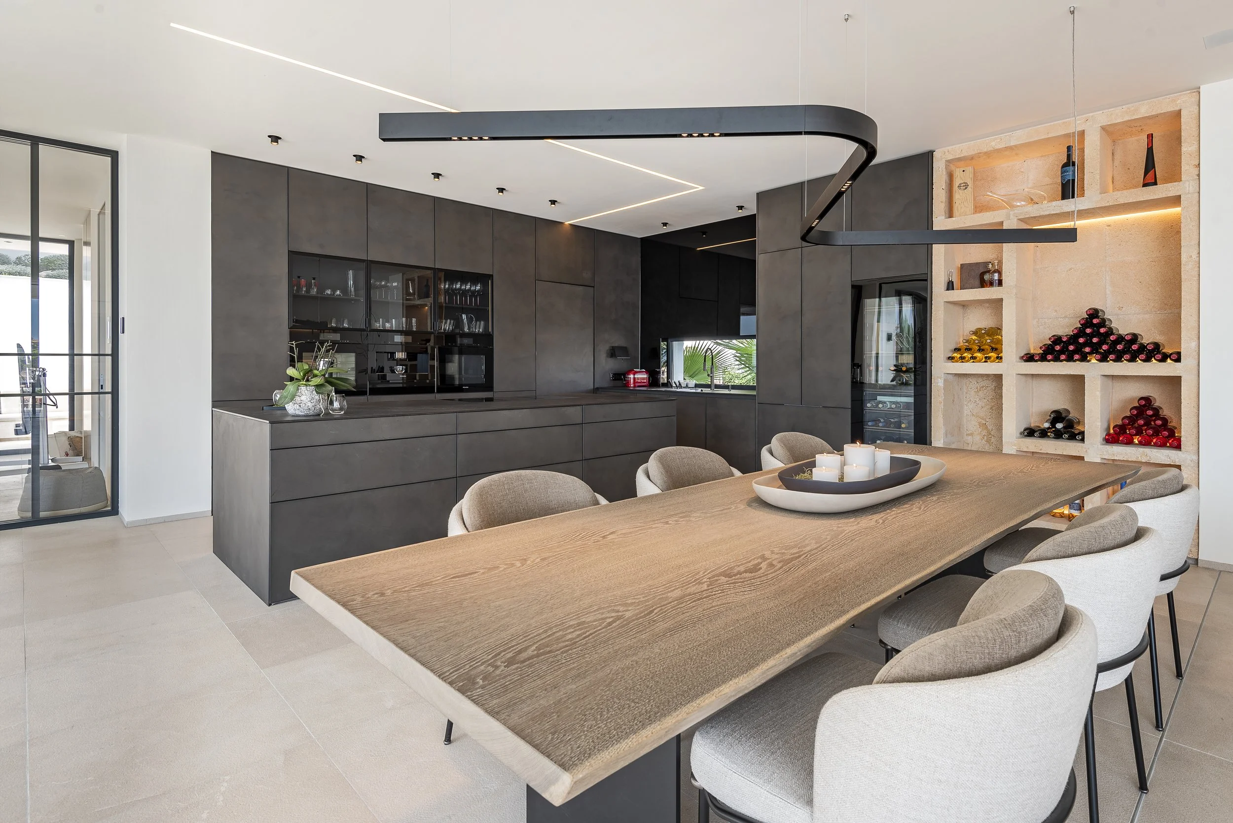 Interior de una casa moderna con cocina y comedor. Mesa de madera con sillas beige, diseño minimalista con electrodomésticos negros, estantería con botellas y vino, y decoración