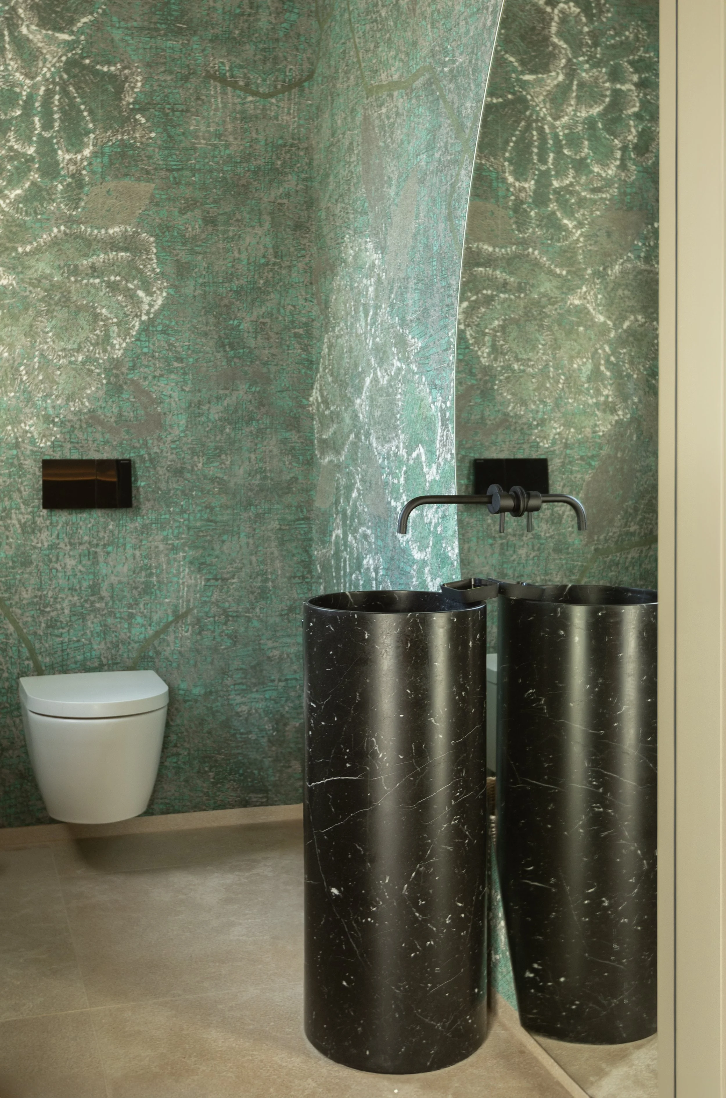 Baño moderno con pared decorativa de mosaico en tonos verdes y grises, sanitarios blancos y lavabos de mármol negro con patas de tubo de metal negro.