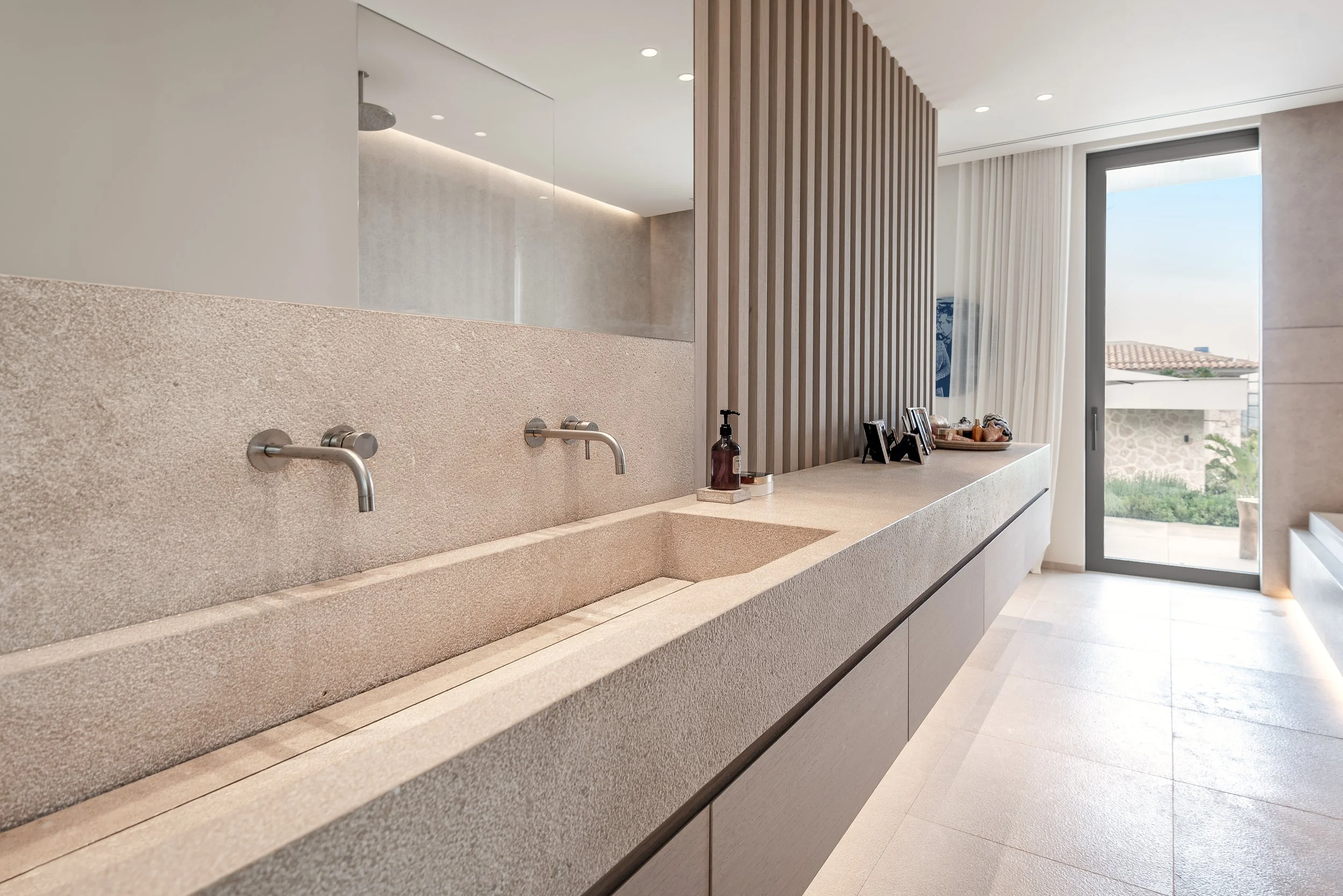Encimera de lavabo de piedra con dos lavabos integrados, accesorios de jabón y fotos en la encimera, con ventanas que permiten la entrada de luz natural y cortinas blancas en el fondo.