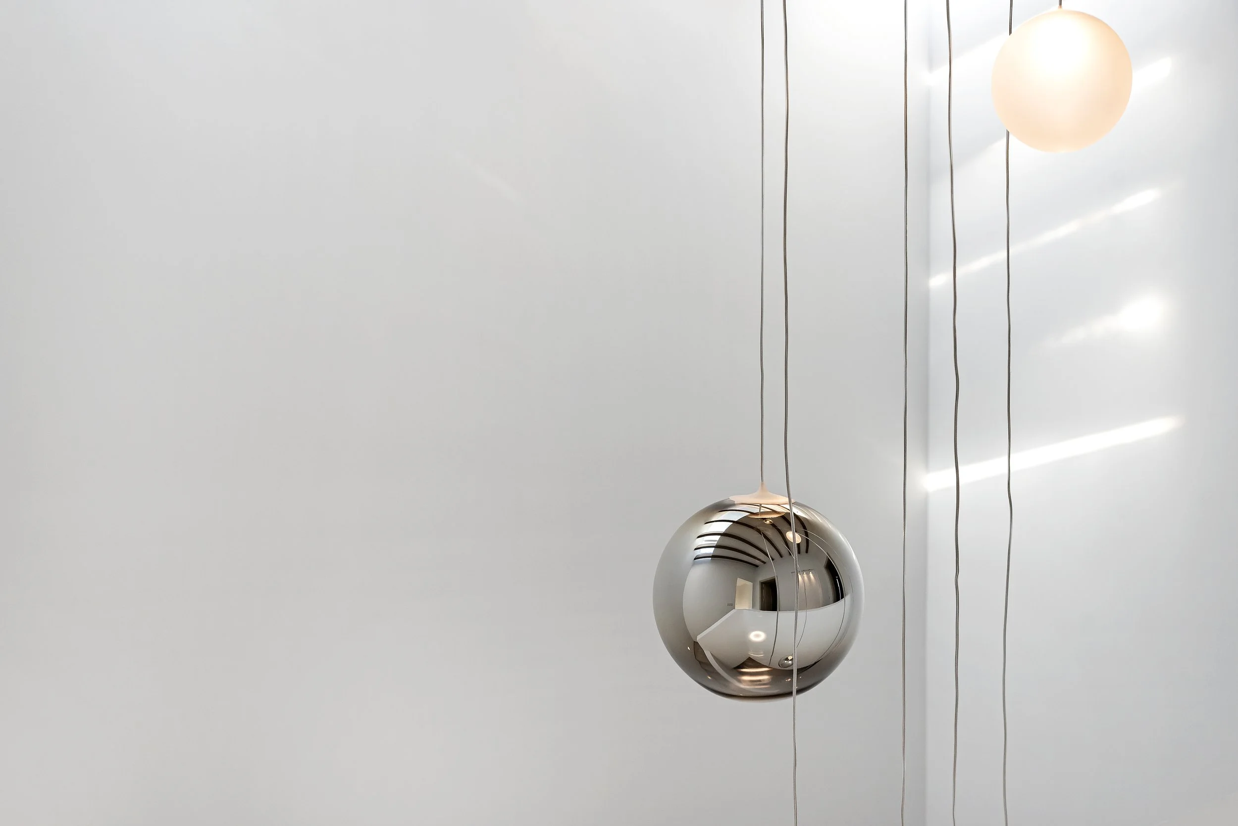 Lámpara colgante moderna de forma esférica de color plateado y brillo metálico, suspendida por cables finos en un ambiente con luz natural y pared blanca.