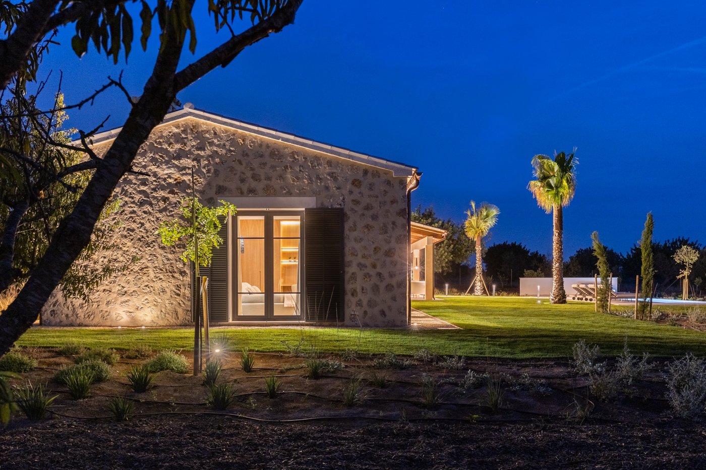 Casa moderna de piedra con iluminación exterior, rodeada de plantas y palmeras, durante la noche.