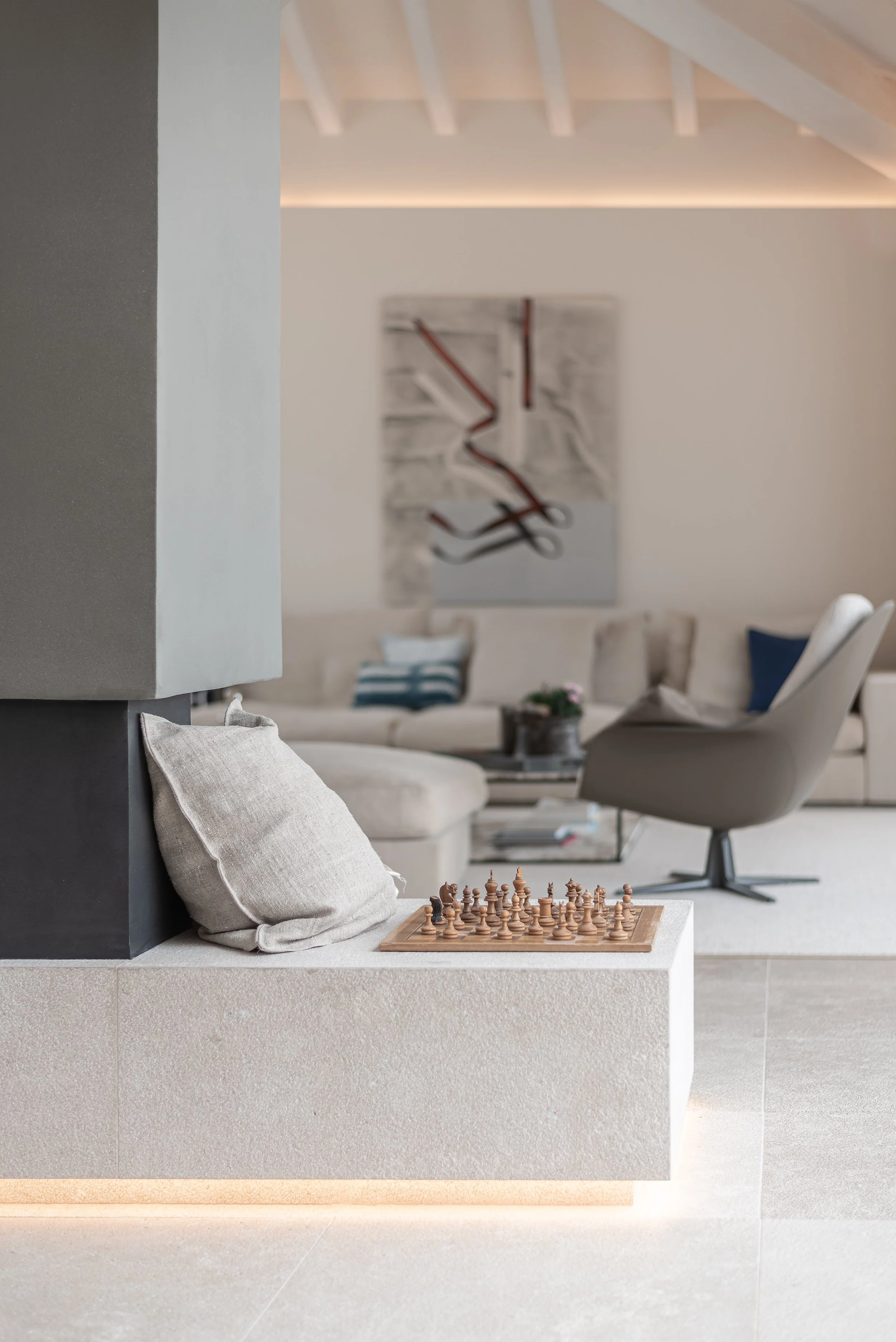 Interior moderno con una silla gris a la derecha, un juego de ajedrez de madera sobre una repisa empotrada en la pared, y un área de descanso con muebles color beige y adornos en tonos neutros en el fondo.
