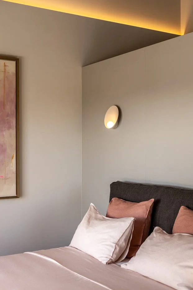 Cama con almohadas rosadas y blancas en una habitación moderna, con luz cálida en la pared y una pintura abstracta.